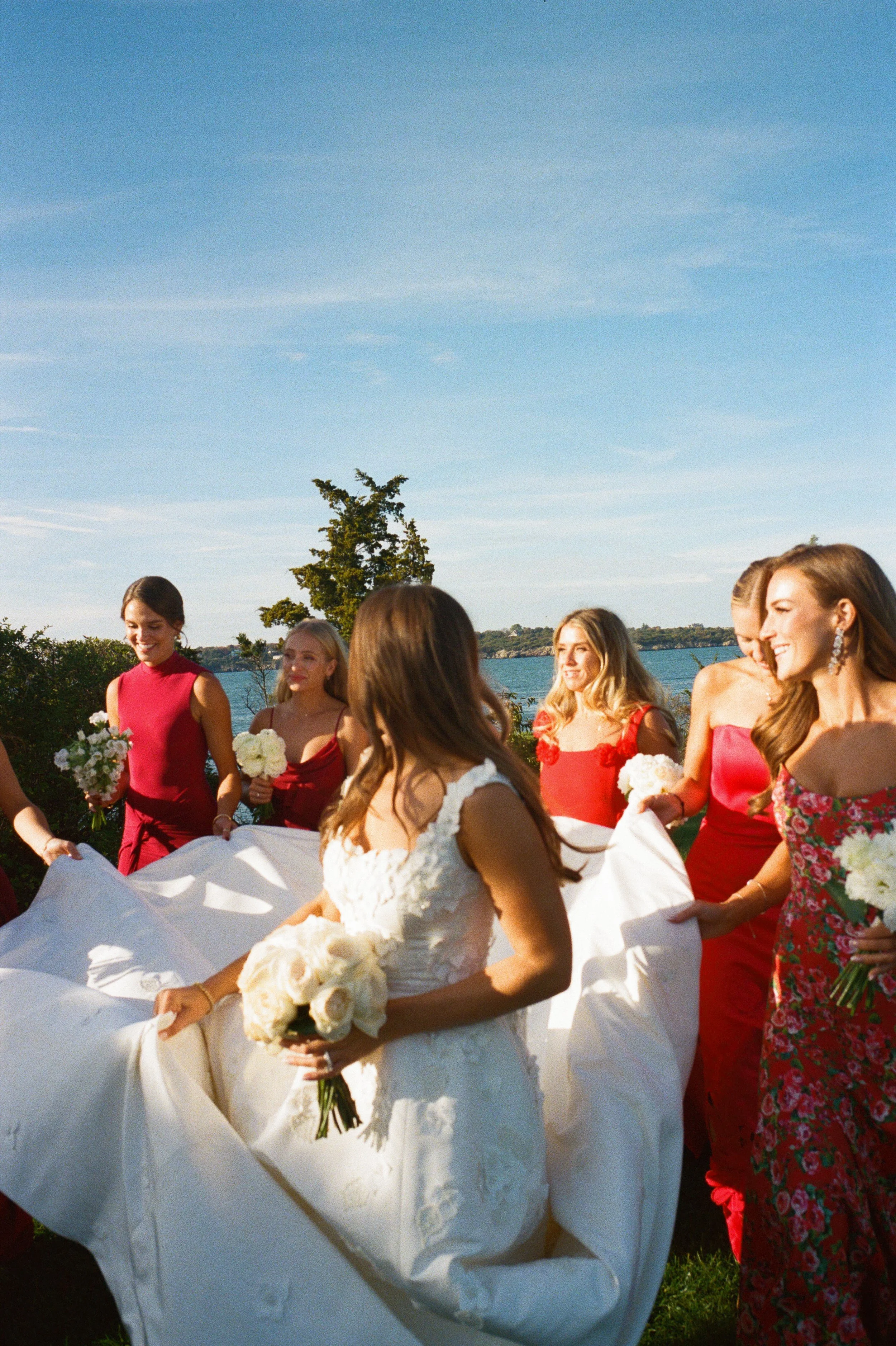 A bride in a white wedding dress holding a bouquet of white roses, surrounded by bridesmaids in red dresses holding bouquets, outdoors near a body of water on a sunny day.