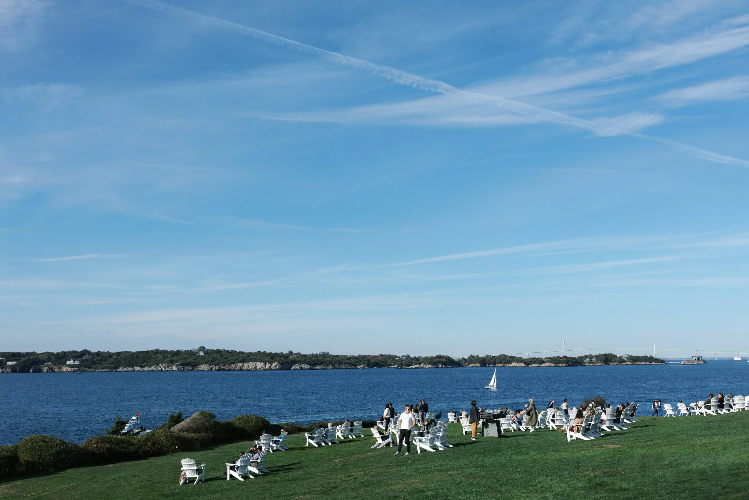 People relaxing on white chairs and standing on a grassy lawn near water, with a sailboat sailing in the distance and a bridge crossing water in the background under a blue sky with wispy clouds.