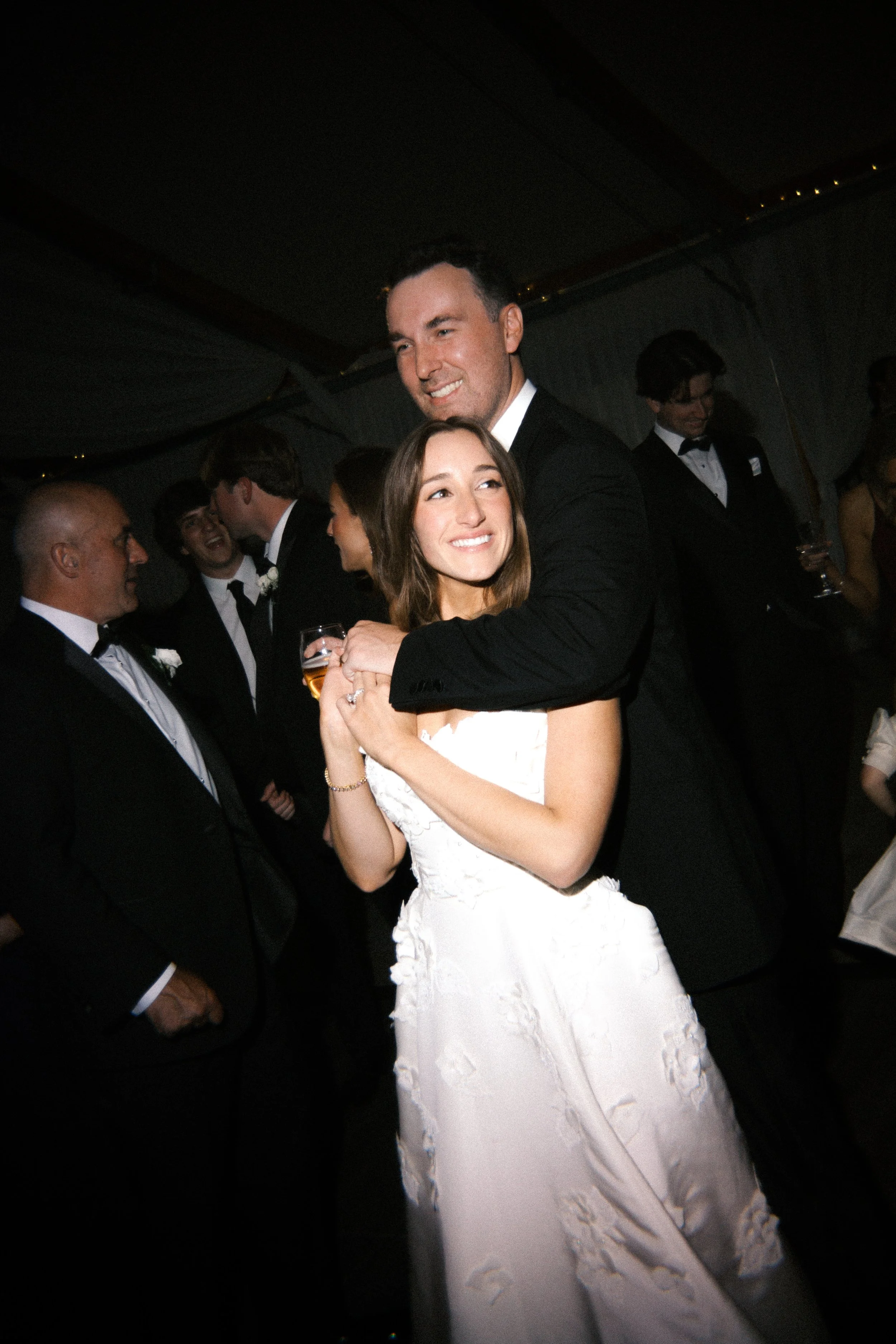 A smiling couple, the man in a tuxedo and the woman in a white wedding dress, dance together at a wedding reception with other guests in the background.