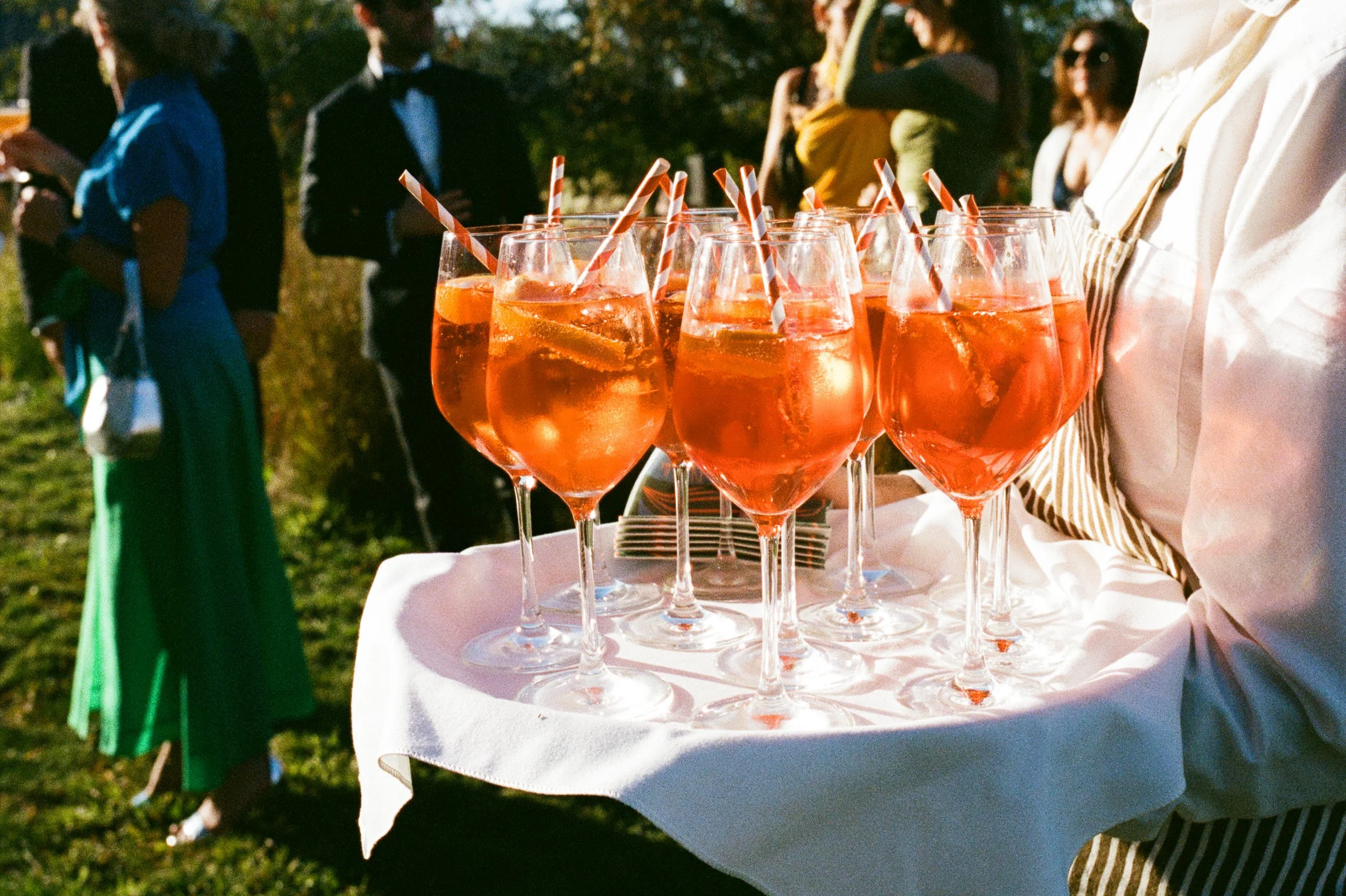 A waiter holding a tray of Aperol spritz cocktails during an outdoor event with people socializing in the background.