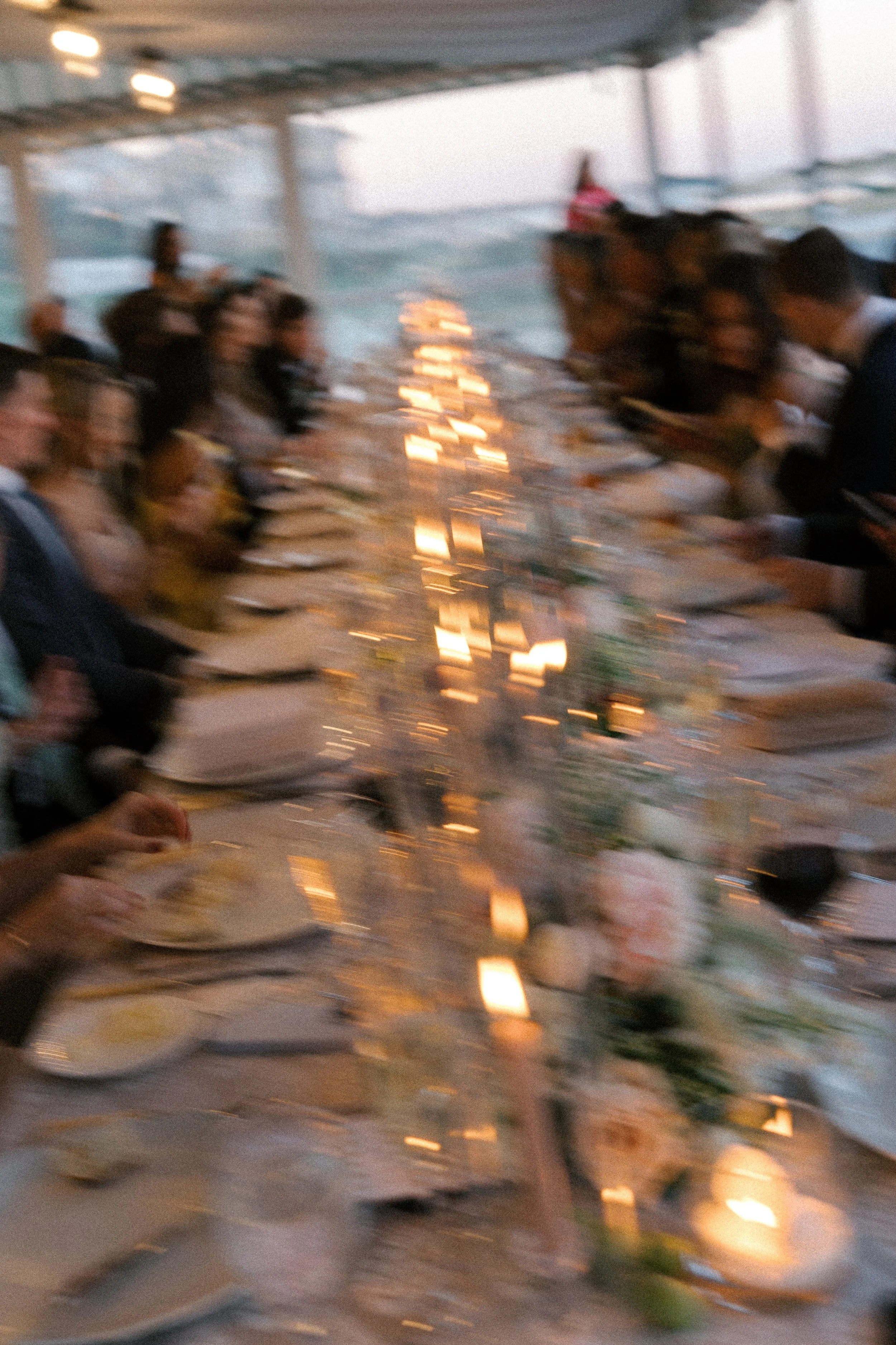 Blurred image of people dining at a long table on a boat, with candles and flowers as decoration, during sunset or dusk.