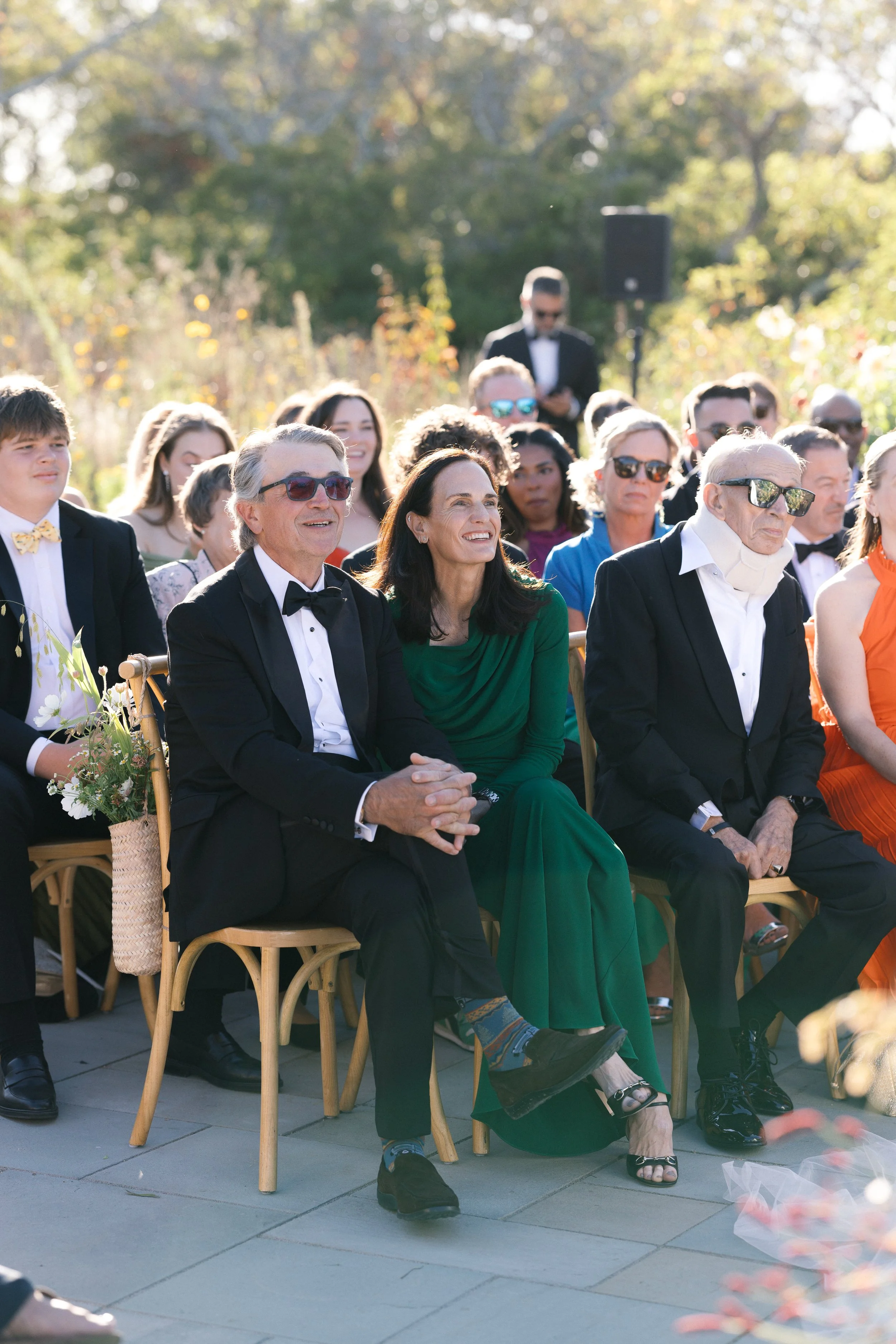 A group of elegantly dressed people sitting outdoors at a formal event, with some wearing suits and sunglasses, on a sunny day with trees in the background.