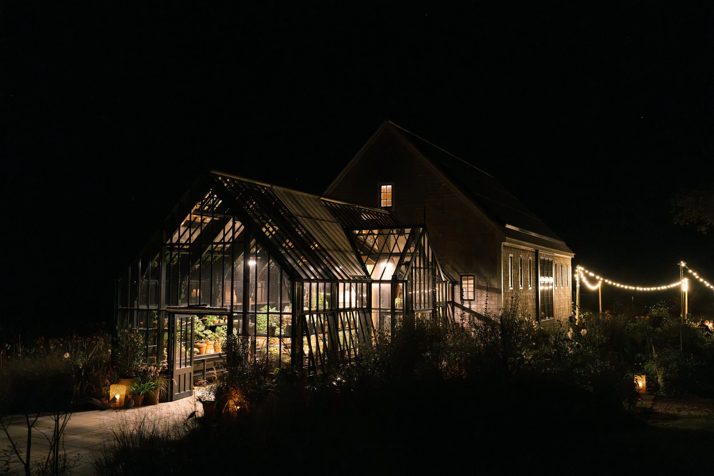 A dark night view of a house with a lit greenhouse attached, illuminated from within, and string lights hanging outside.