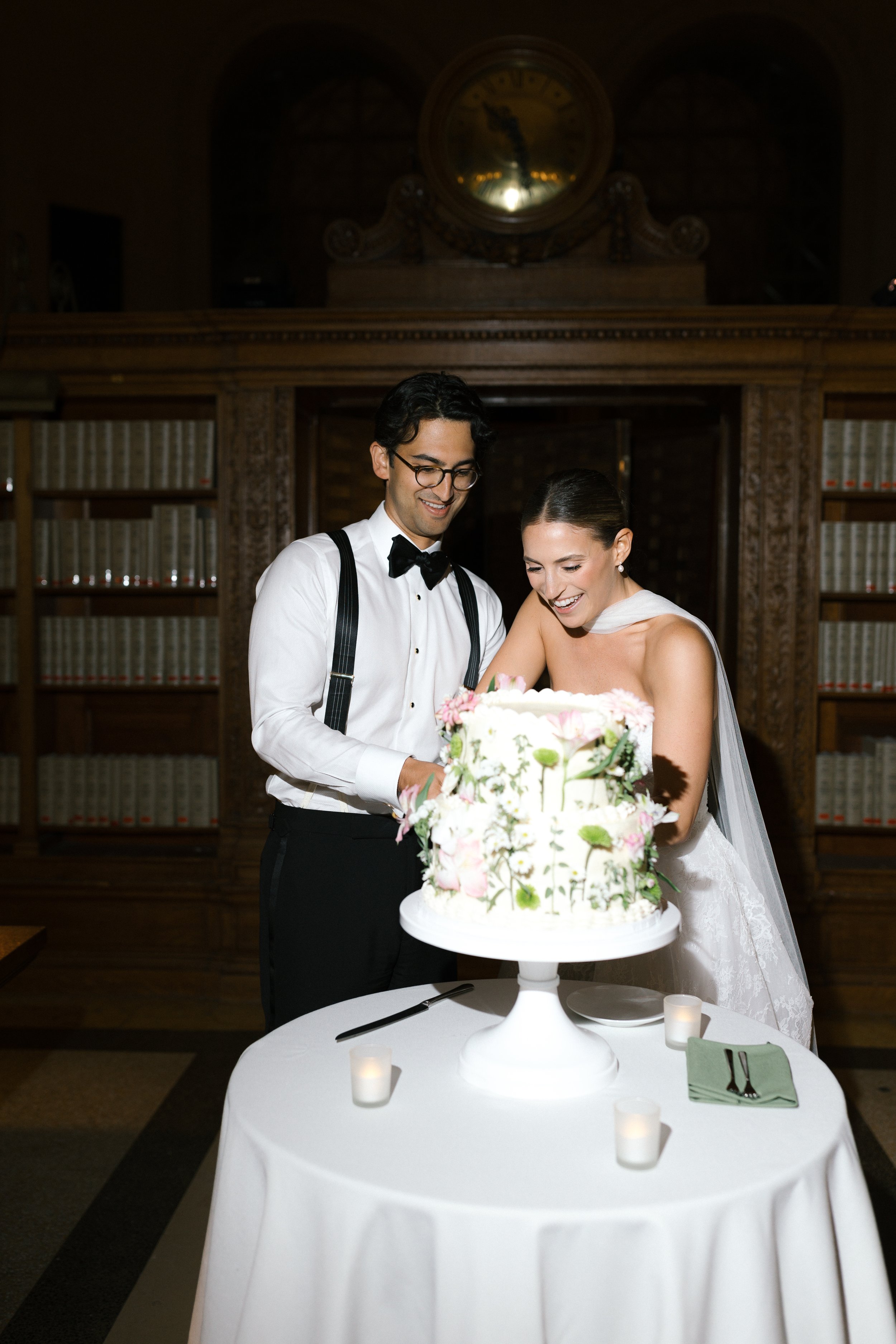 Happy bride and groom cutting a wedding cake together at their wedding reception in a wood-paneled room with bookshelves in the background.