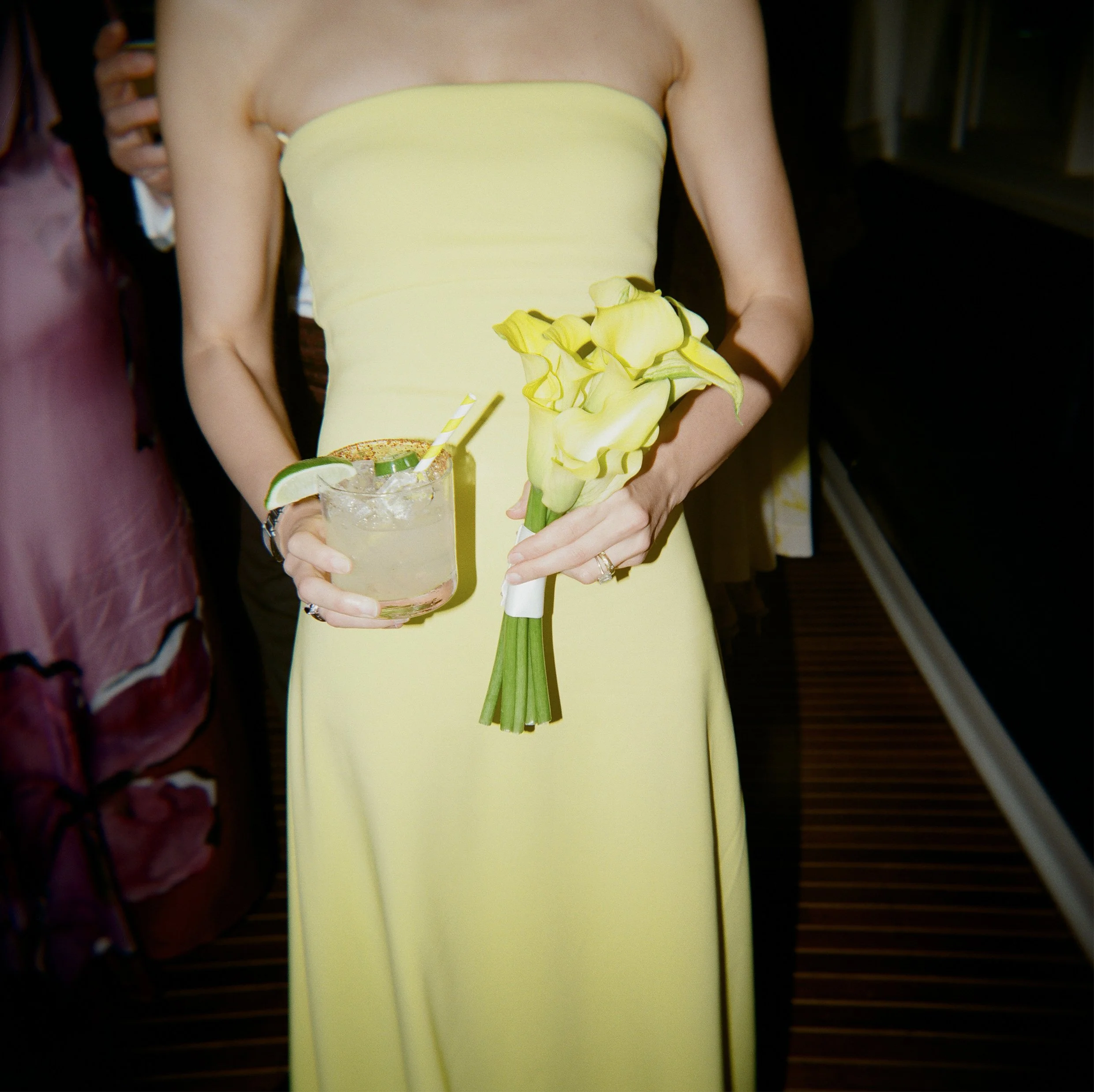 A woman wearing a yellow strapless dress holding a bouquet of yellow calla lilies and a cocktail glass with lime wedge and ice.