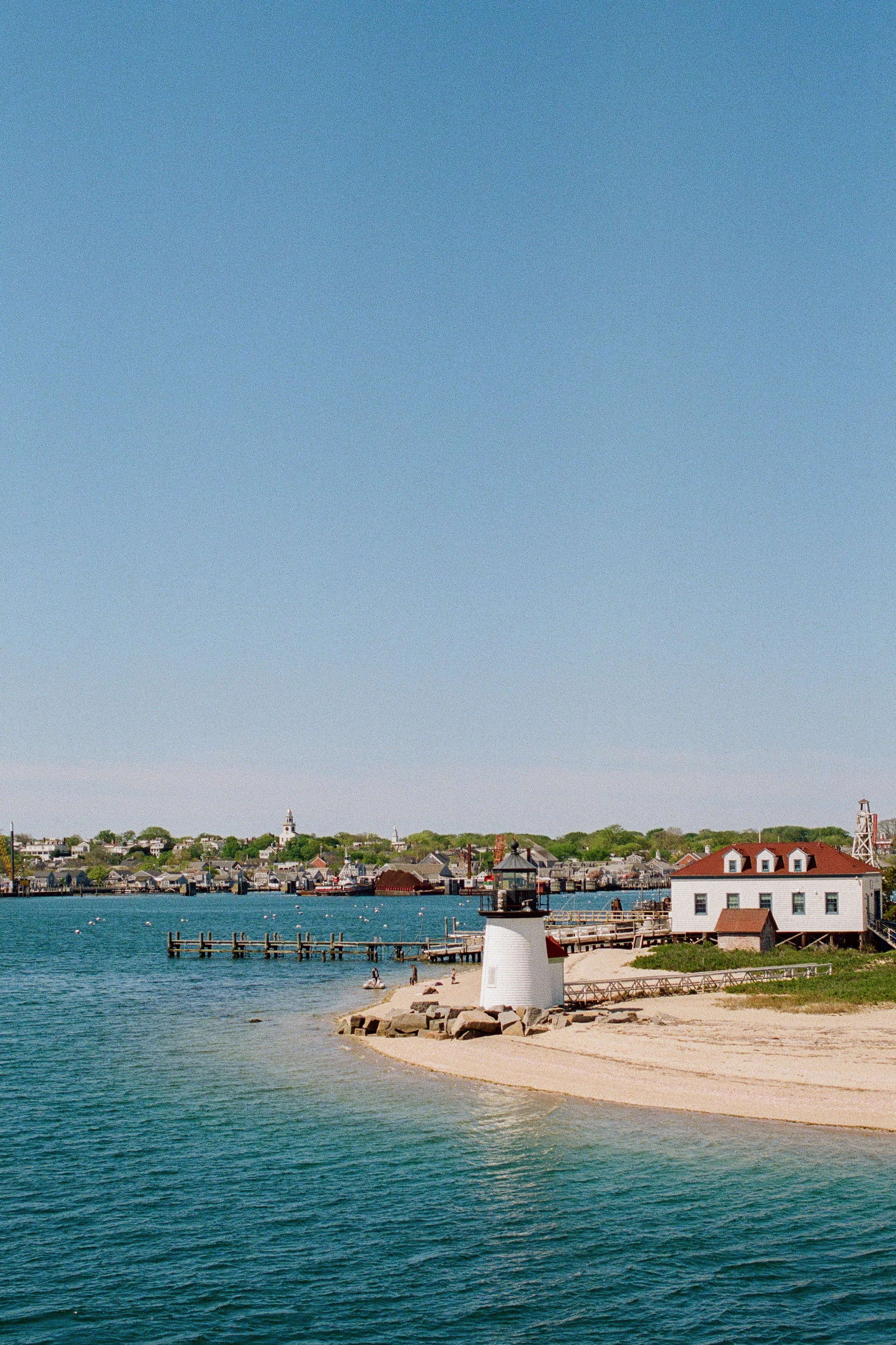 A lighthouse on a sandy beach with a small building nearby, with a pier extending into calm water and a town in the background under a clear blue sky.