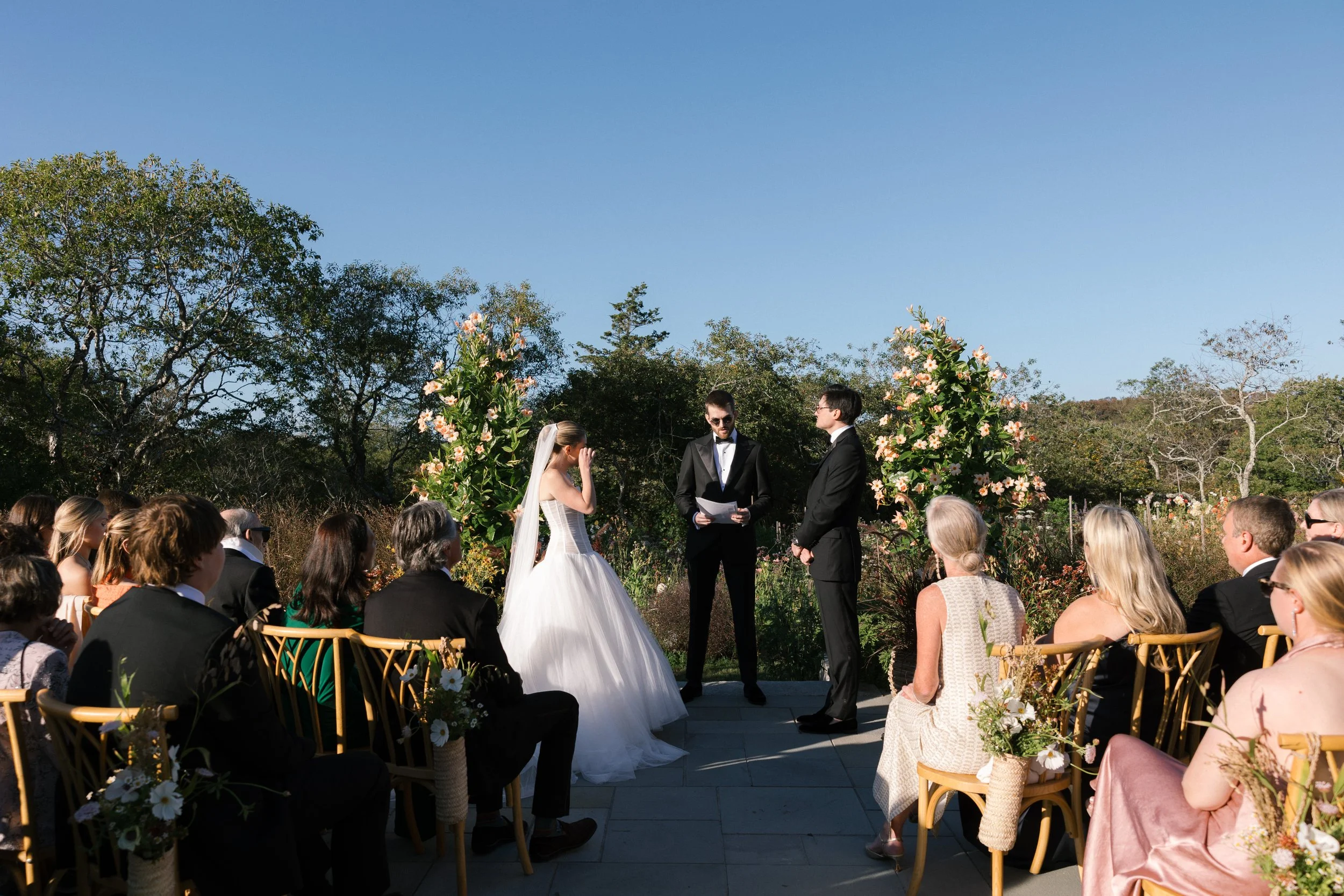 Outdoor wedding ceremony with a bride and groom exchanging vows, officiant reading, guests seated in front, floral arrangements, and greenery background under clear blue sky.