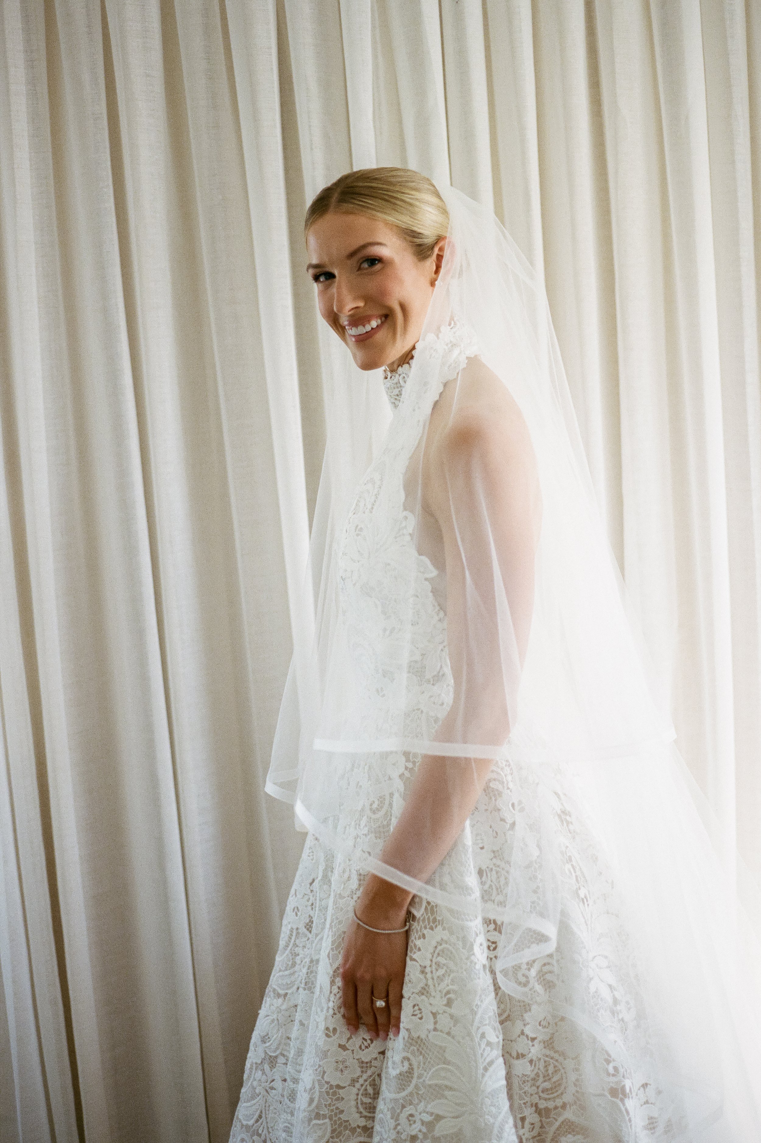 A smiling bride with blonde hair in a bridal gown with lace details, standing next to a cream-colored curtain.
