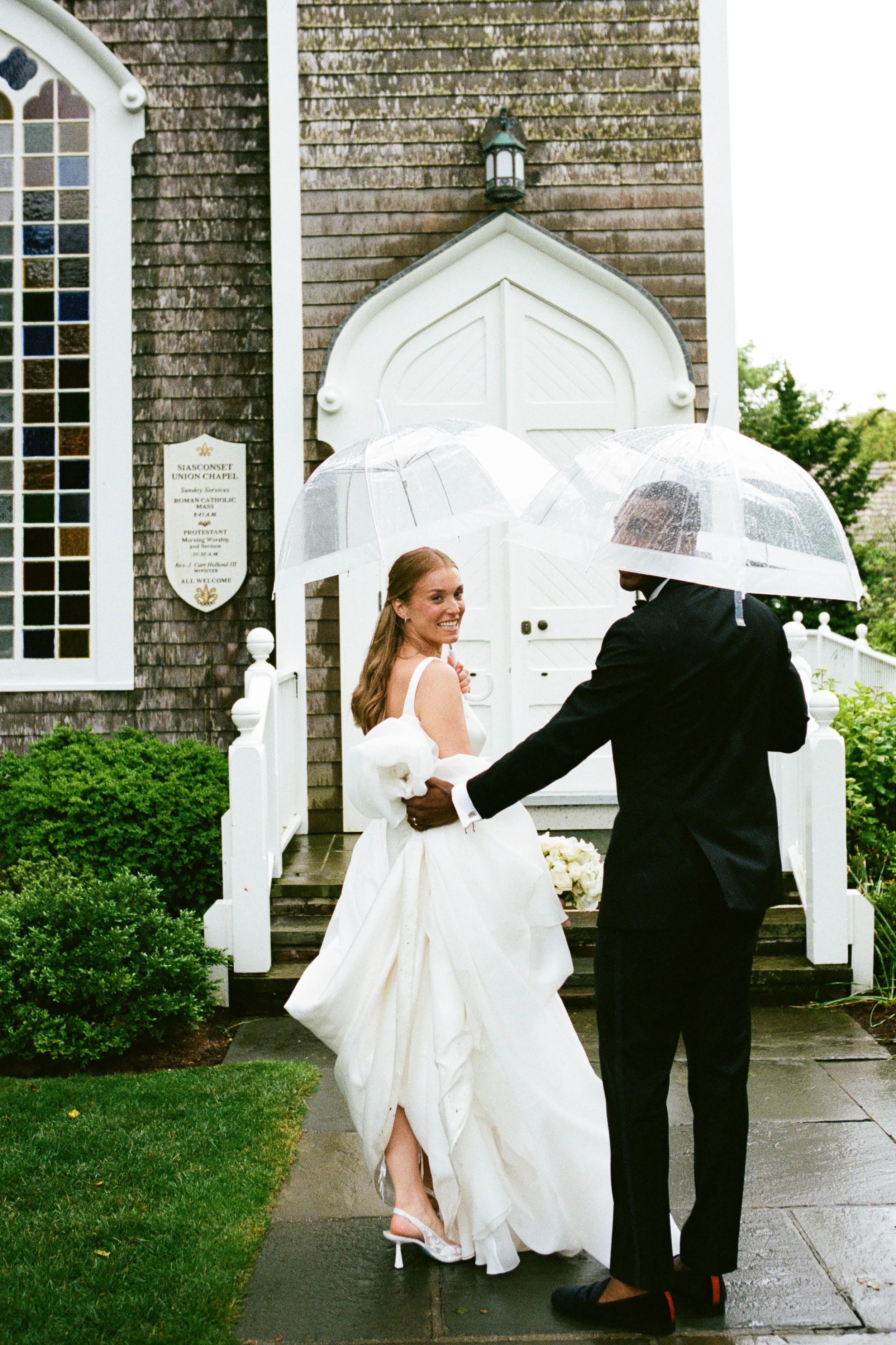 A bride in a white wedding gown and a groom in a black suit holding umbrellas walking on a wet sidewalk outside a church.