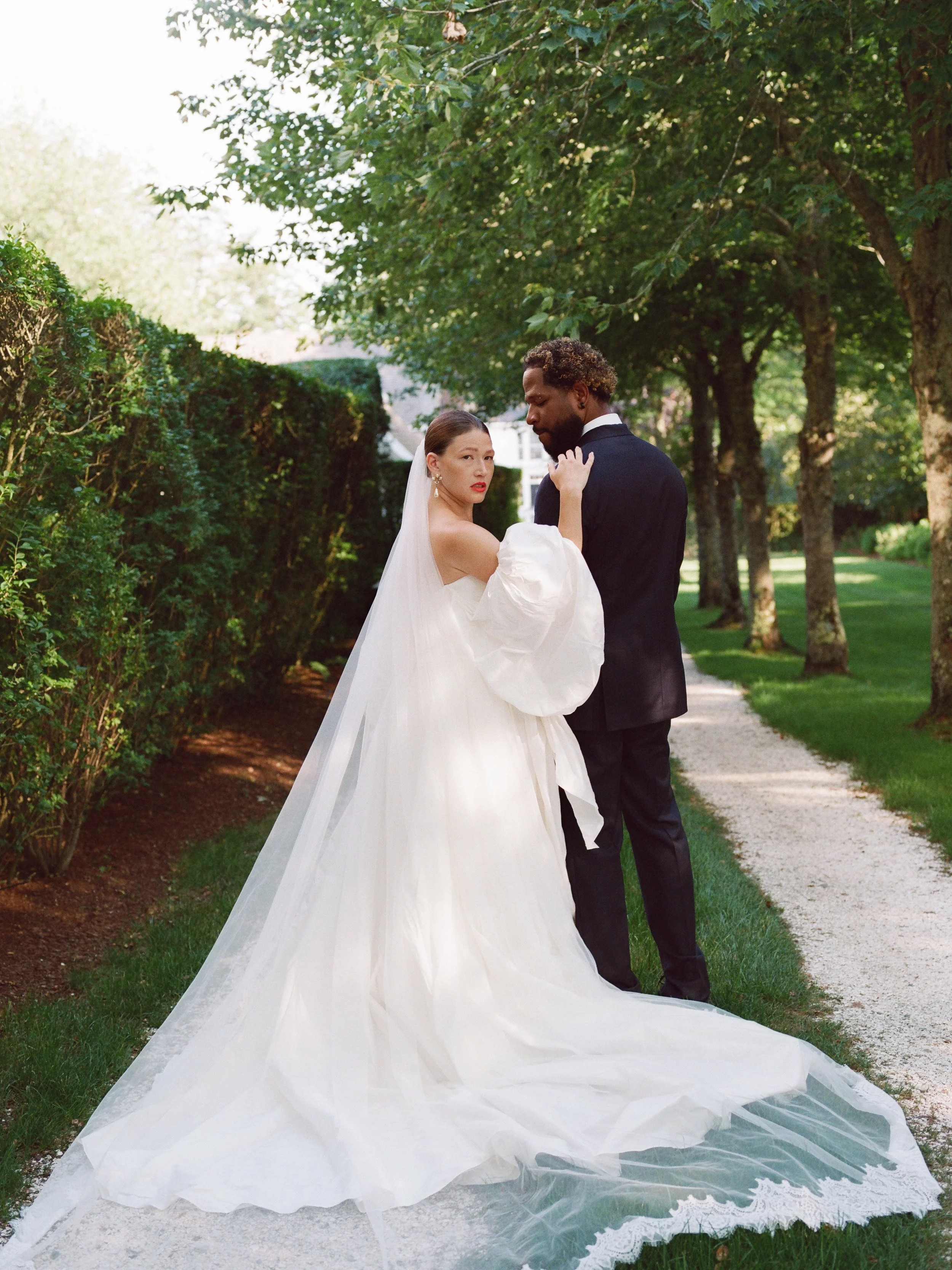 A bride and groom standing on a garden path lined with trees, with the bride in a white wedding gown and veil, and the groom in a black suit, embracing outdoors.