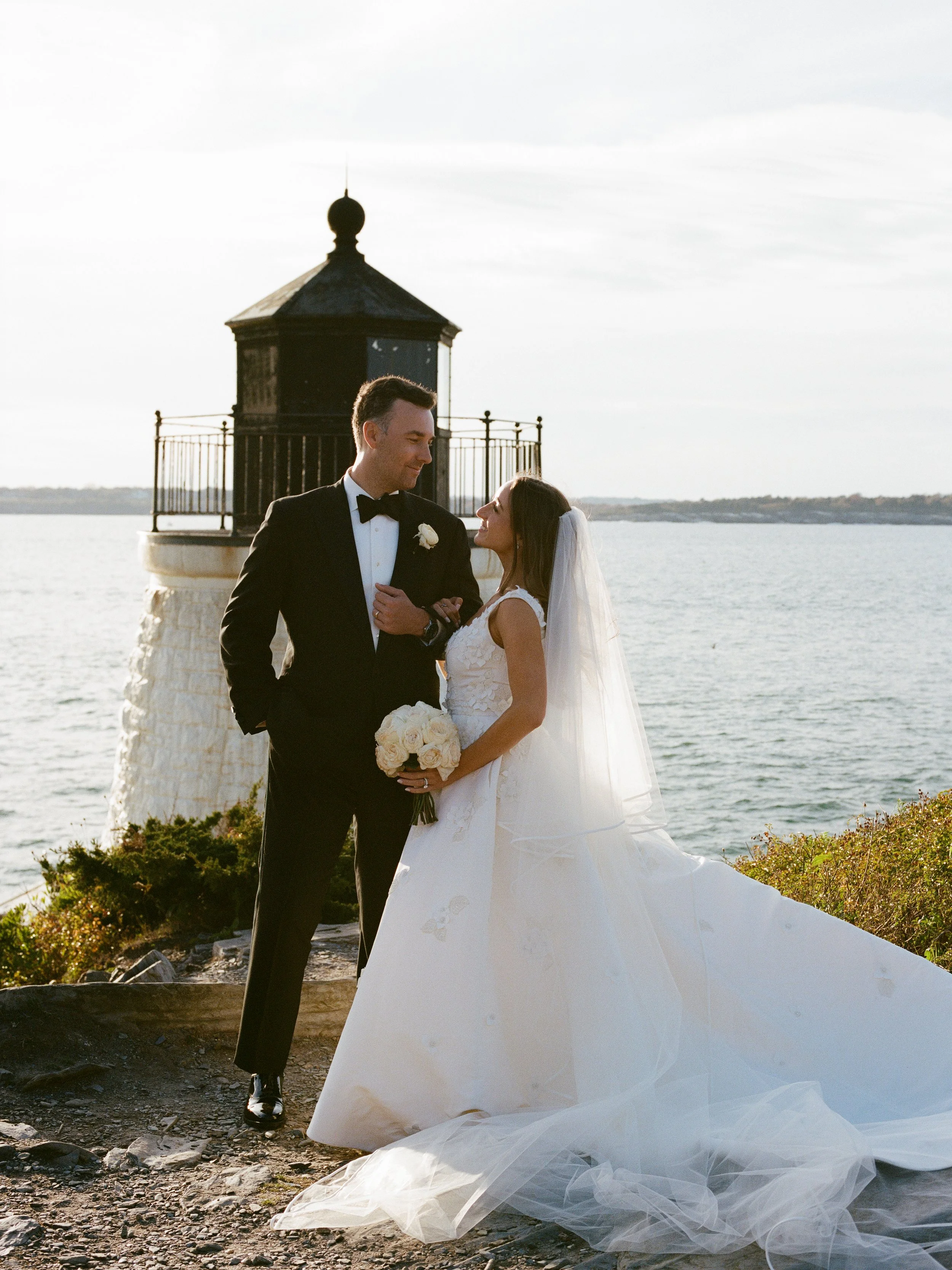 A bride and groom standing together near a lighthouse by the water, smiling and looking at each other during their wedding photoshoot.