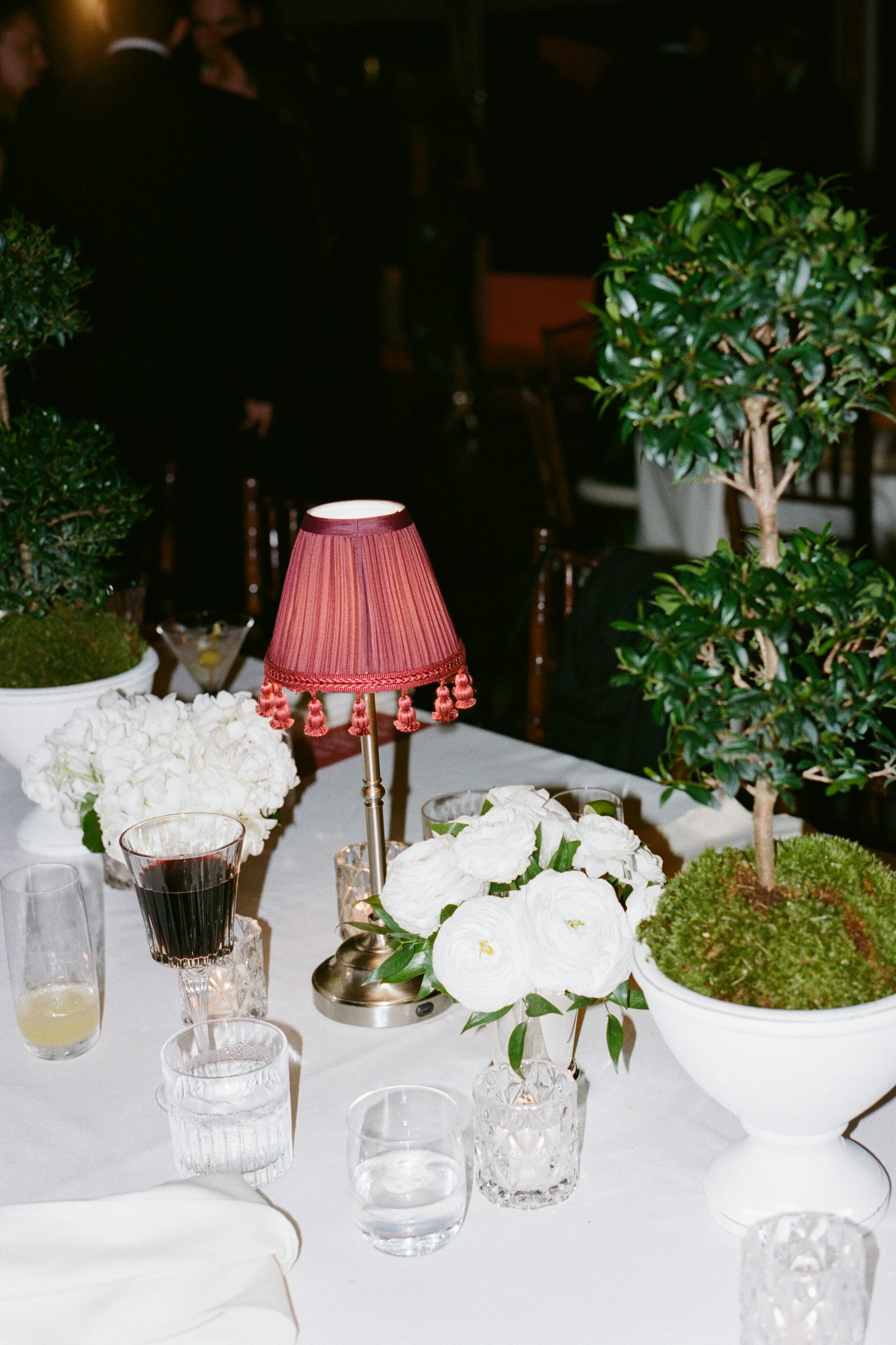 A table decorated with white flowers, a pink lampshade, and potted green plants at a formal event or restaurant.