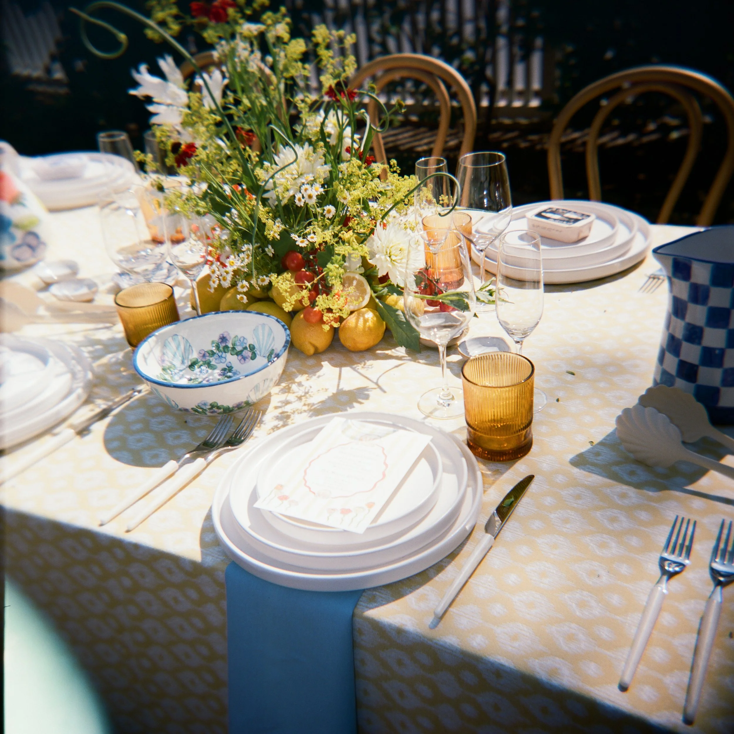 A decorated outdoor table set for a meal, with white plates, glasses, cutlery, a floral centerpiece, and a blue and white checkered pitcher.