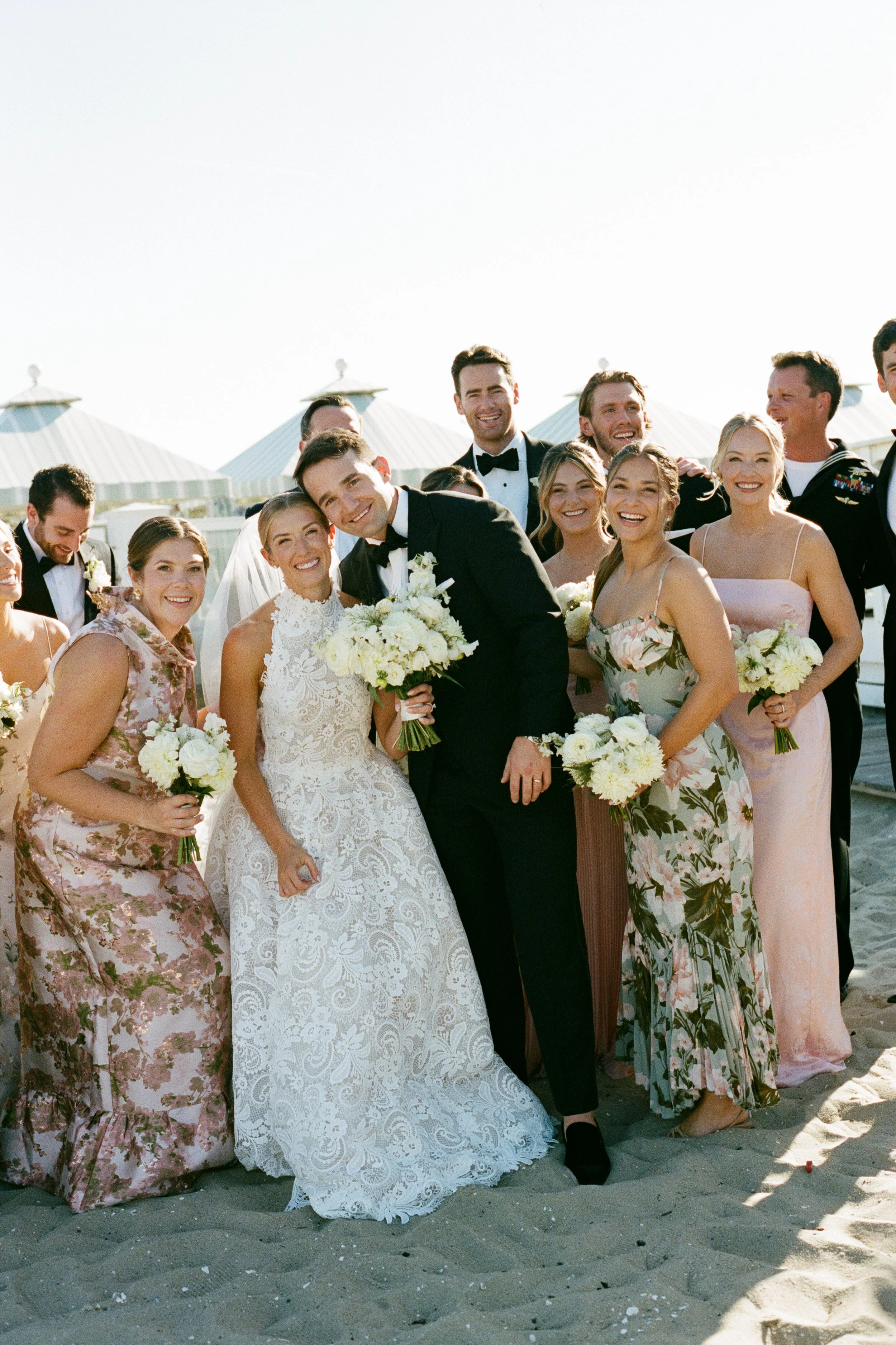 A group of people dressed in formal attire, including a bride in a white wedding gown and a groom in a black tuxedo, posing on the beach with a background of beach huts.