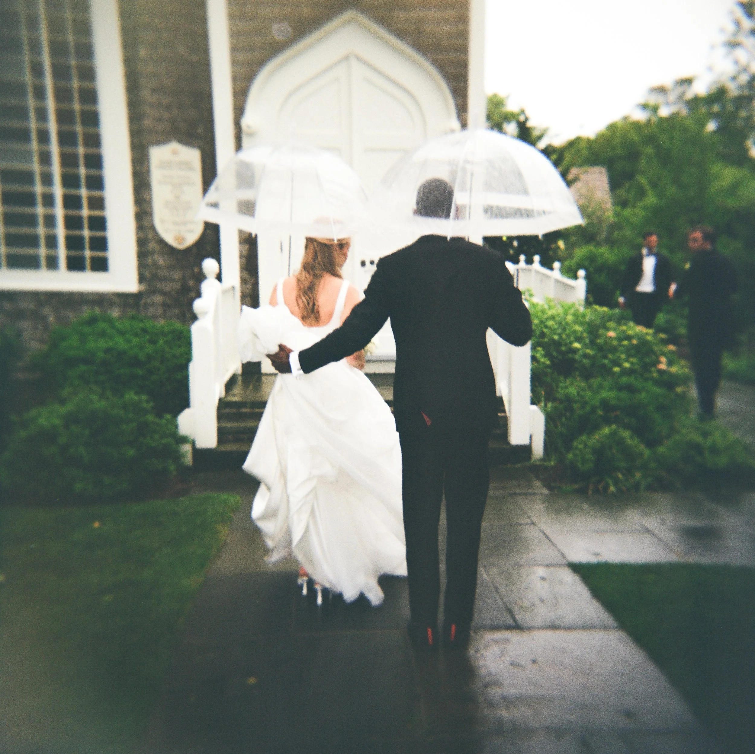 A bride in a white wedding dress and a groom in a black suit walk toward a church entrance under a rainy sky, sharing clear umbrellas, with other guests in the background.
