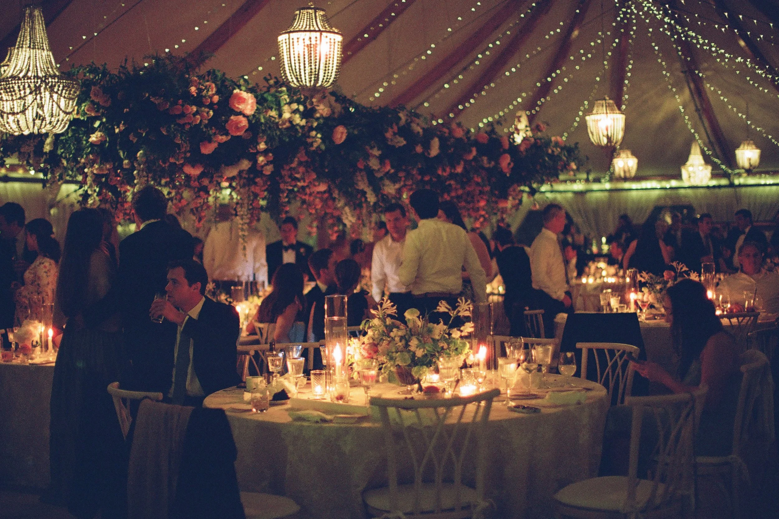 A wedding reception or formal event inside a decorated tent with hanging chandeliers, string lights, and floral arrangements. Guests are seated at tables with candles, and some are standing and chatting.