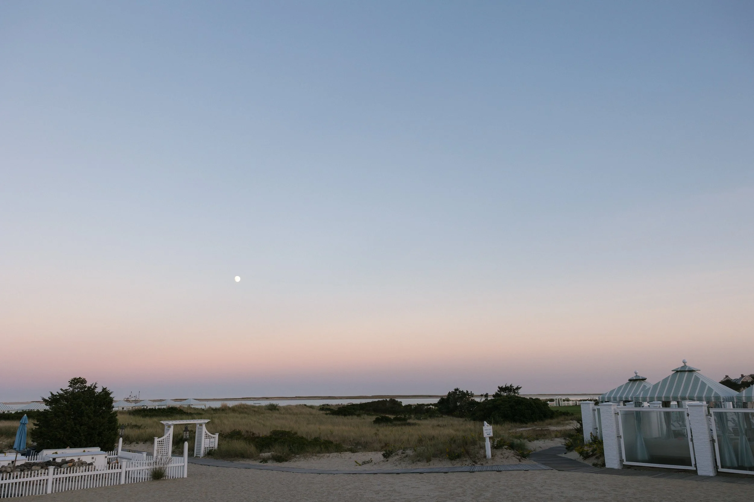 Beach scene at dawn with a pastel-colored sky, the moon visible, white picket fences, sandy path, and beachside cabanas with striped awnings.