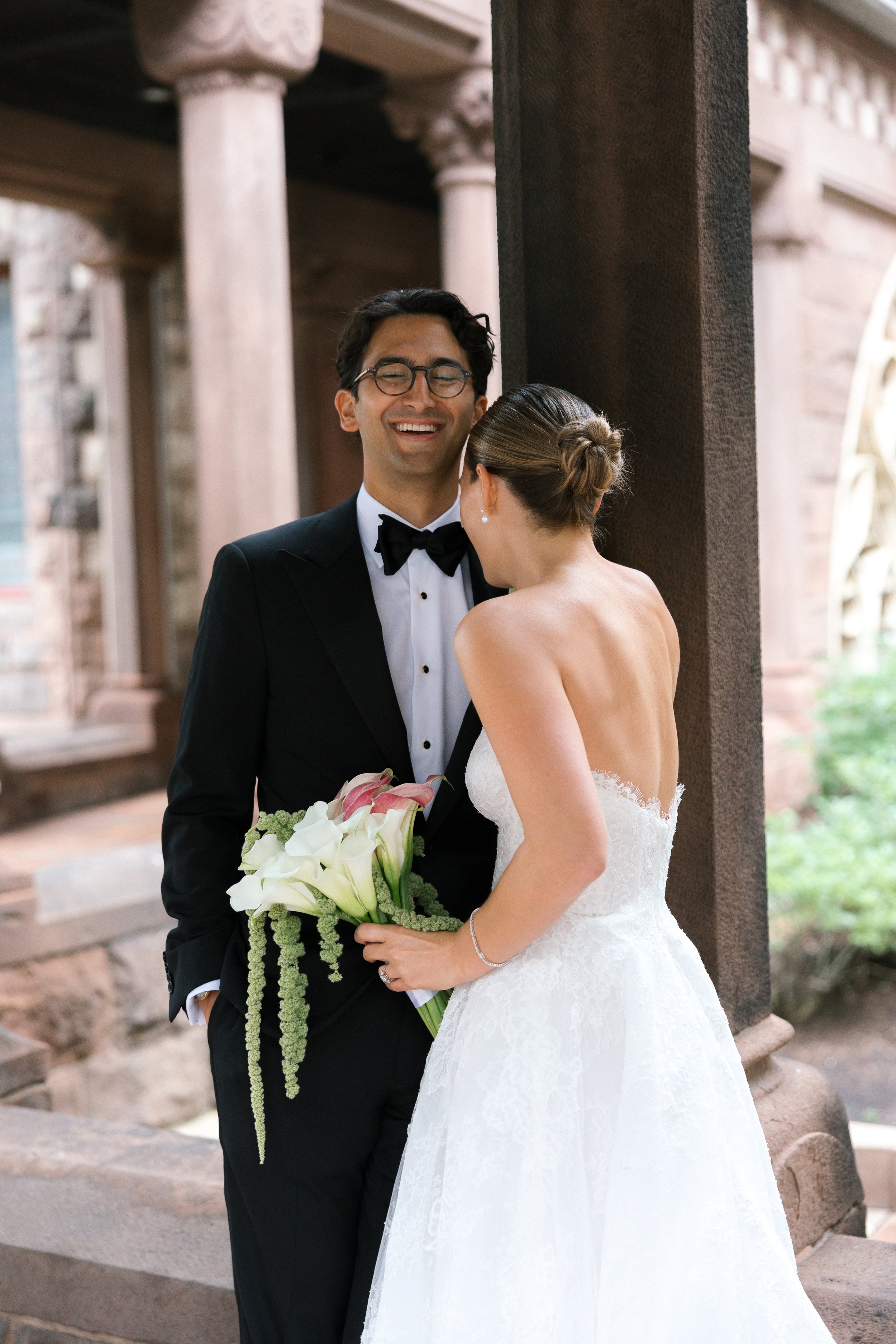 A bride and groom standing close together outdoors, with the bride holding a bouquet of flowers. The groom is wearing a tuxedo and glasses; the bride is in a wedding dress with her hair in an updo.