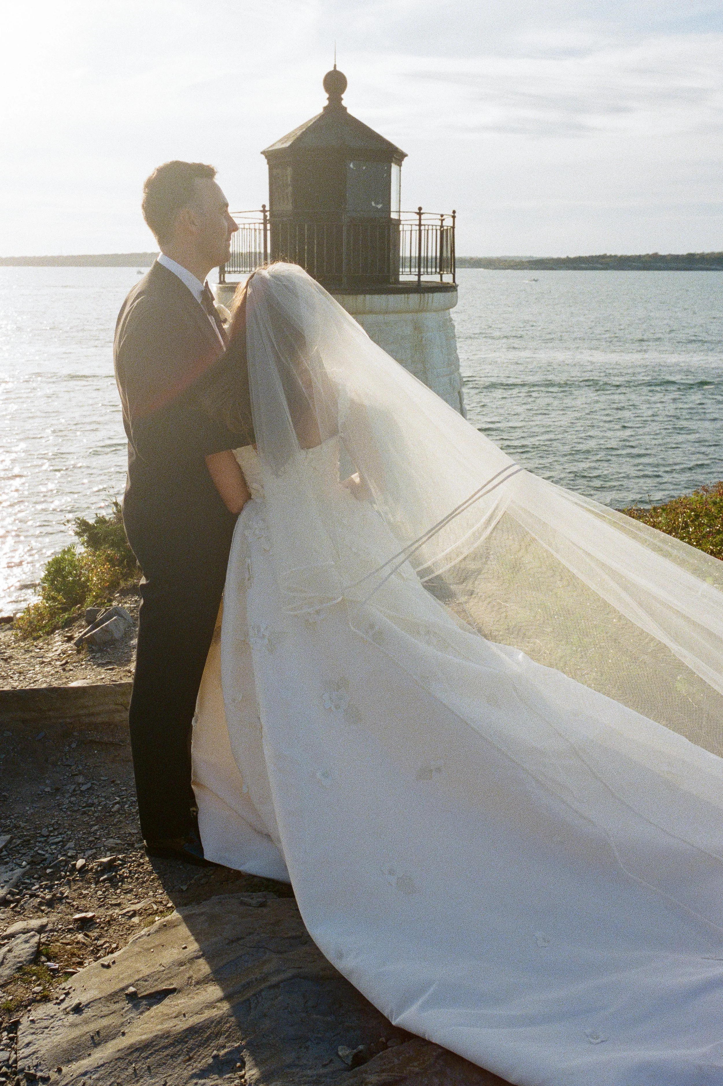 A bride and groom standing by the water during sunset, with a lighthouse in the background.