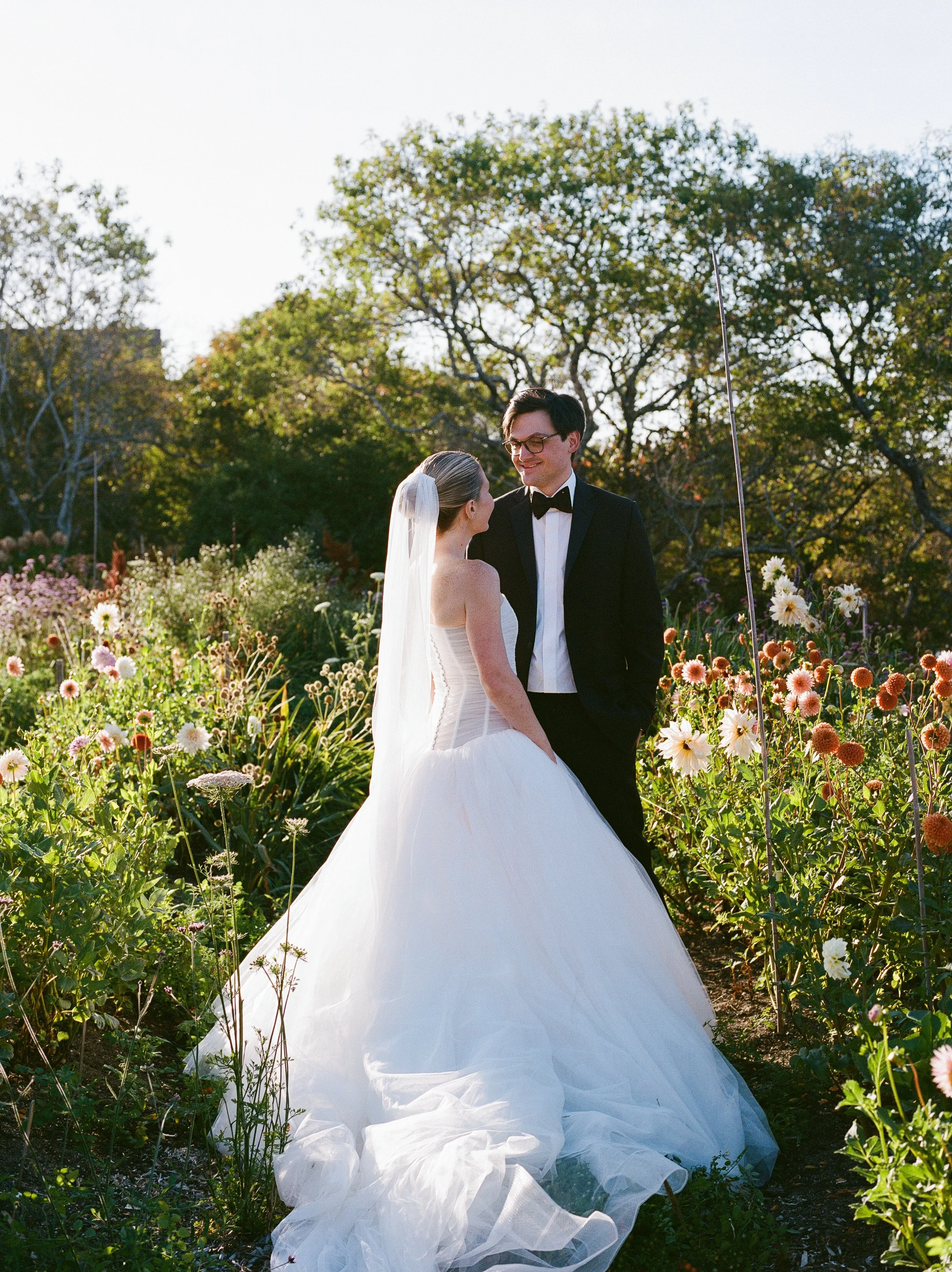 A bride and groom on their wedding day in a garden filled with colorful flowers, with trees and a bright sky in the background.