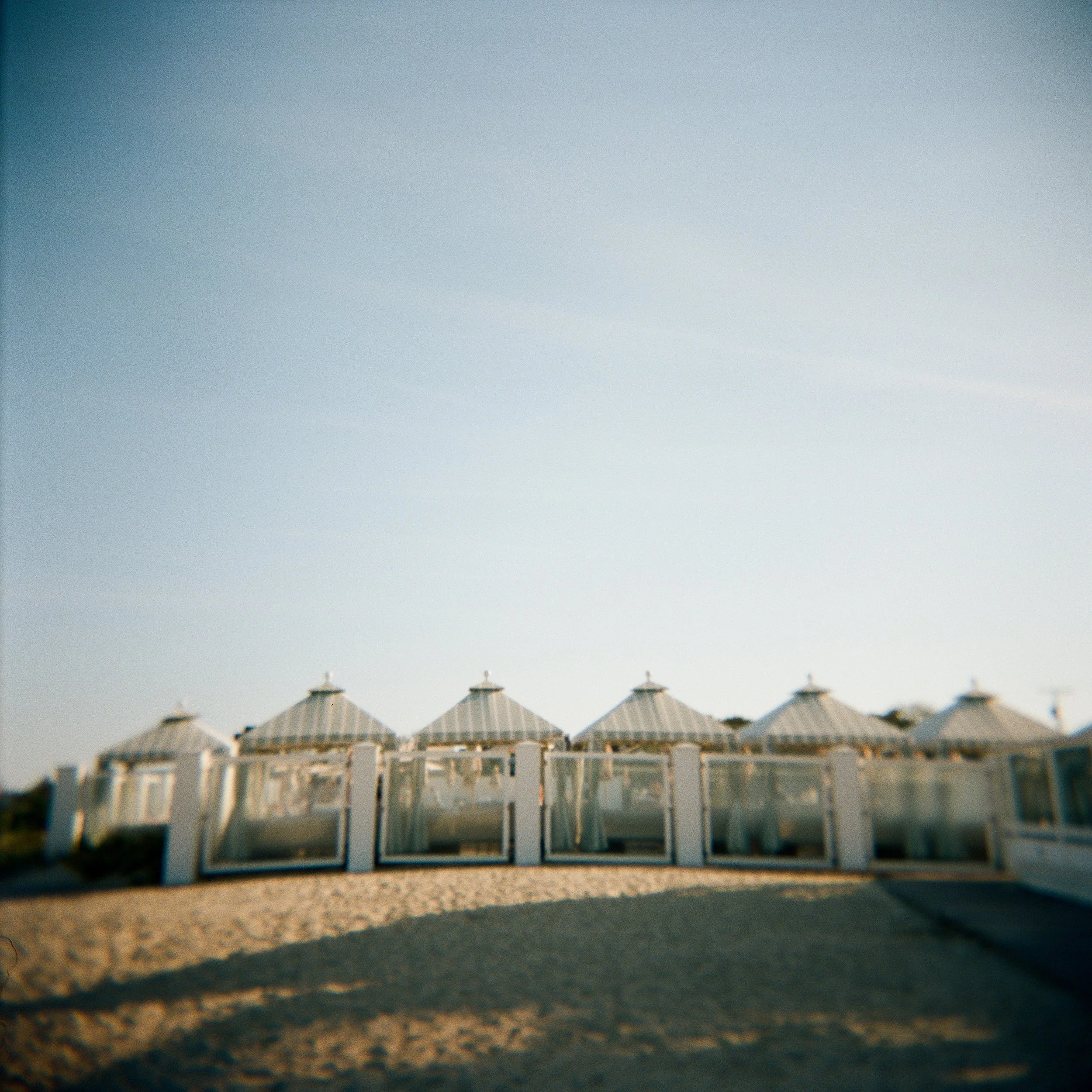 Row of white beach cabanas with pointed roofs on a sandy beach under a clear sky.