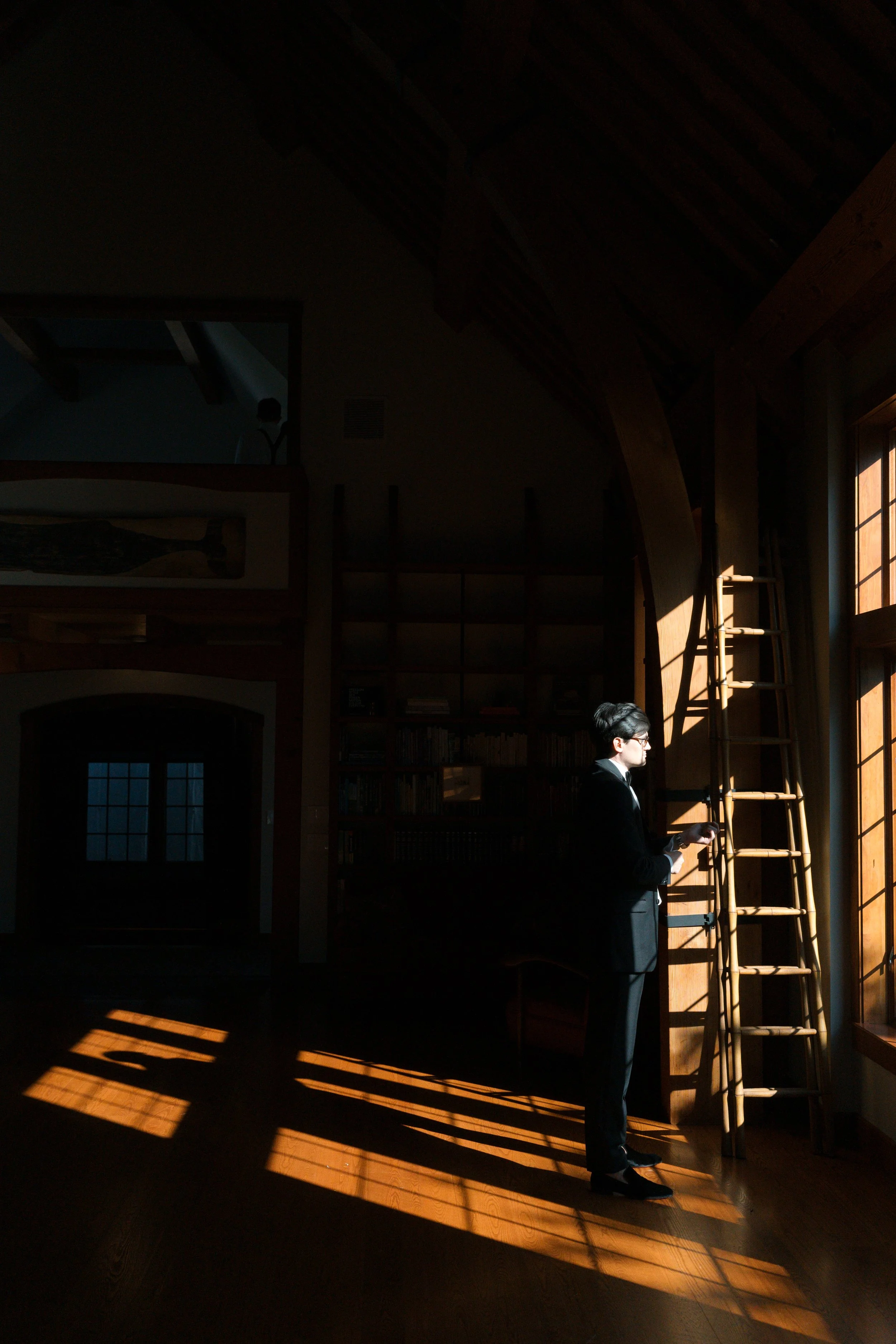A woman in a black suit and glasses is standing in a dimly lit room by a large window, reading a book. Sunlight streams through the window, casting shadows on the wooden floor and a ladder leaning against the wall.