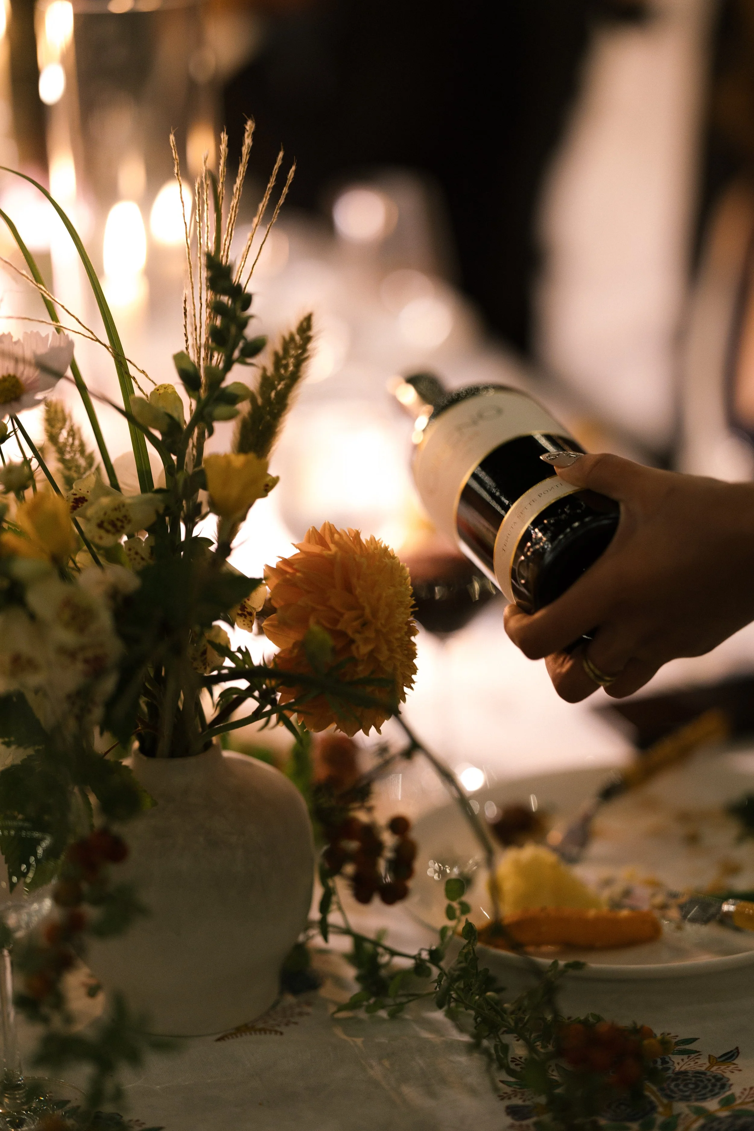A hand holding a black and beige bottle of sparkling wine over a table with a floral centerpiece, wine glasses, and a partially eaten cake, in a dimly lit setting.