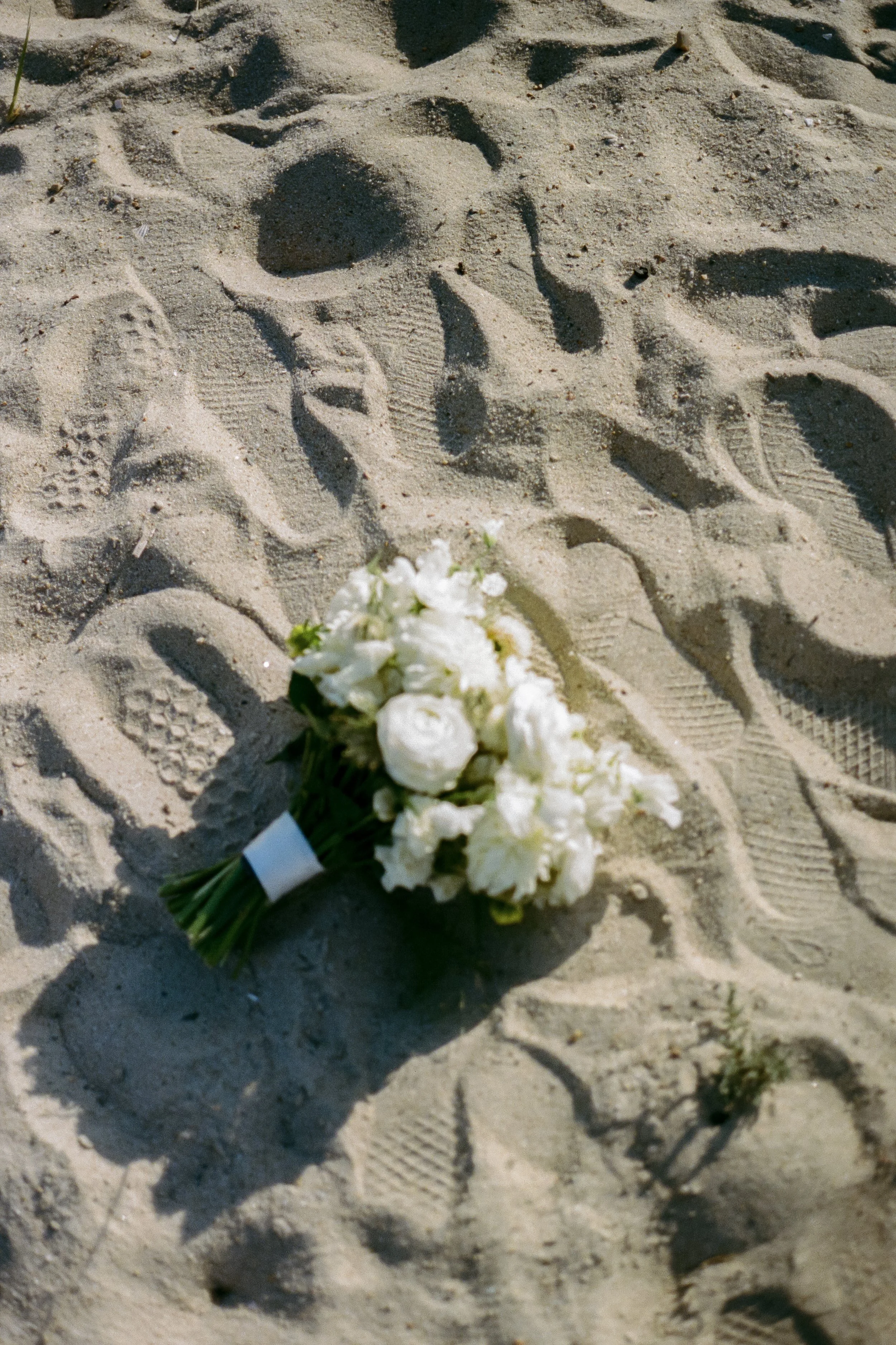 A bouquet of white flowers lying on sandy ground with numerous footprints.