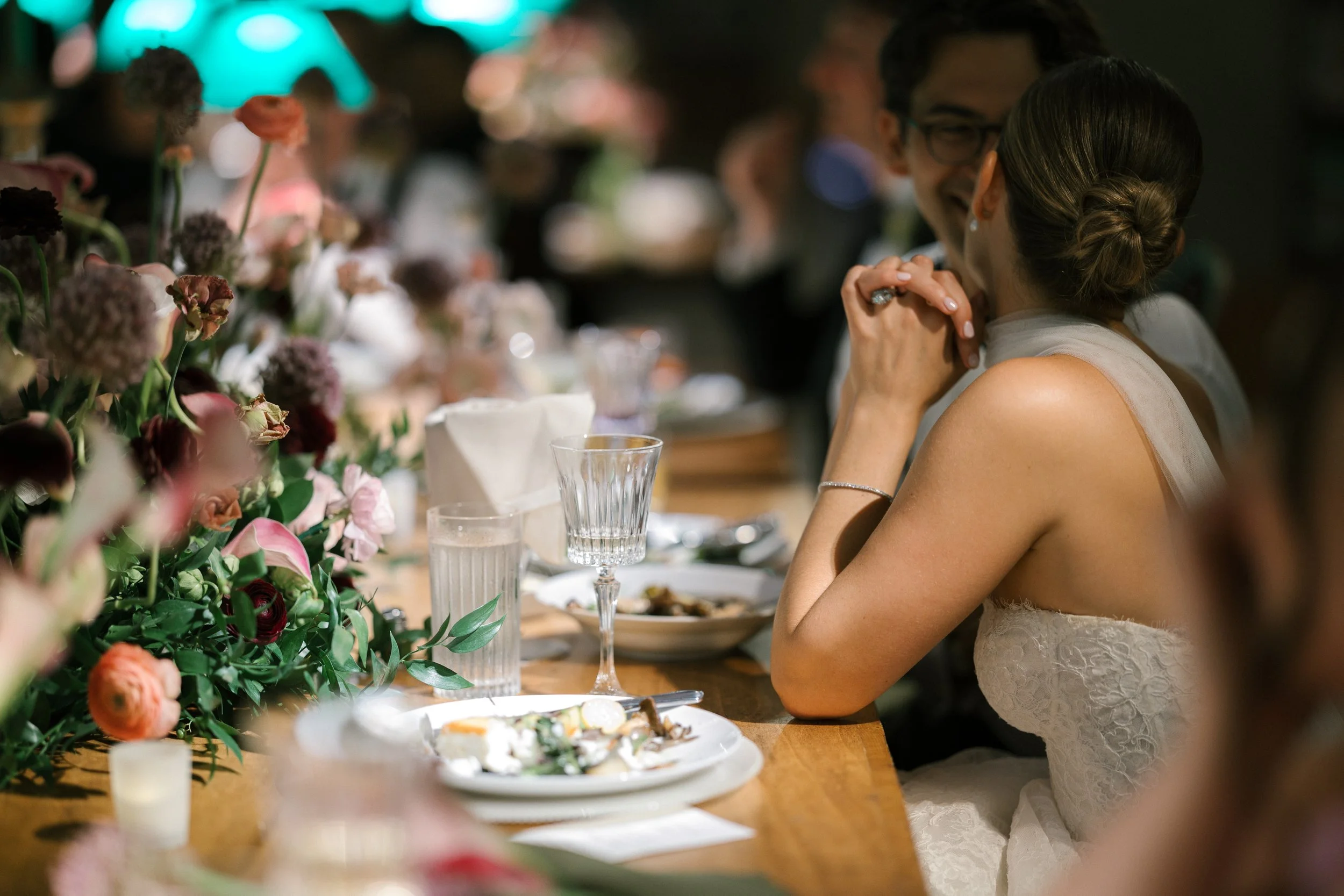 A woman in a white lace dress with her hair in a bun and a man in glasses sitting across a dinner table decorated with flowers, glassware, and plates during an elegant event or wedding reception.