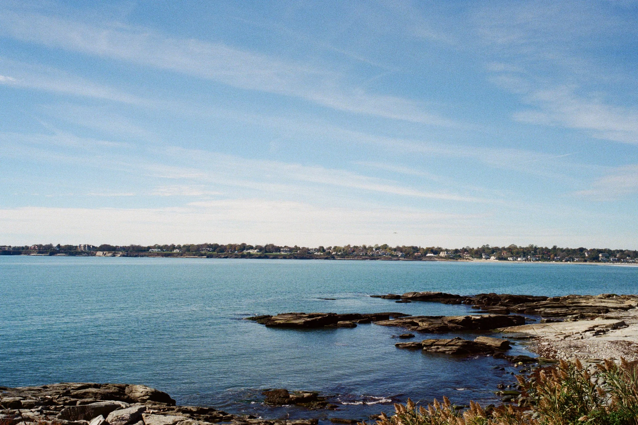 A coastal scene with calm blue water, rocky shoreline, and a distant landmass with houses under a partly cloudy sky.