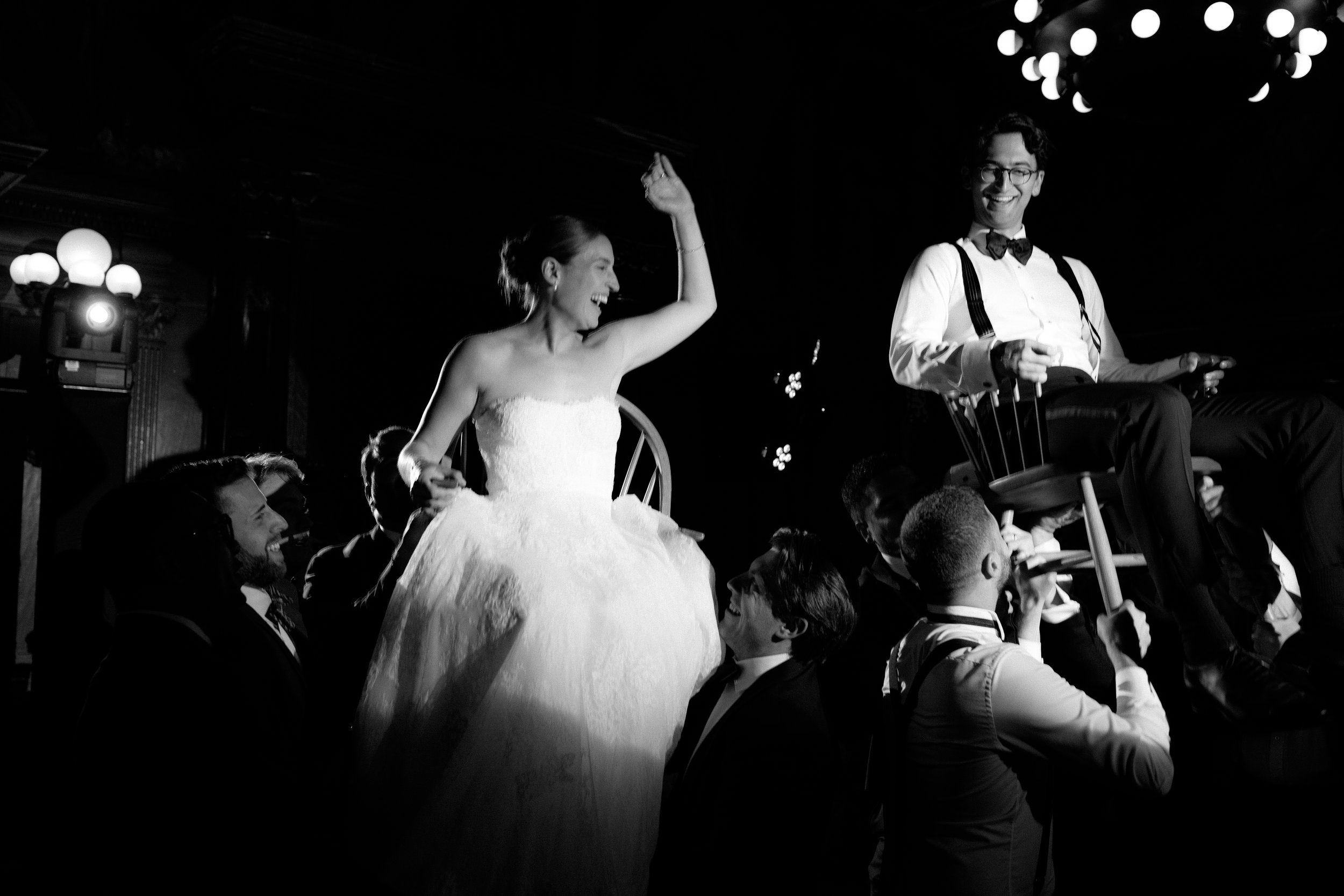 A bride in a wedding dress sitting on a chair being lifted on people's shoulders during a wedding celebration. A groom in a bow tie and suspenders sitting on a chair nearby is smiling.