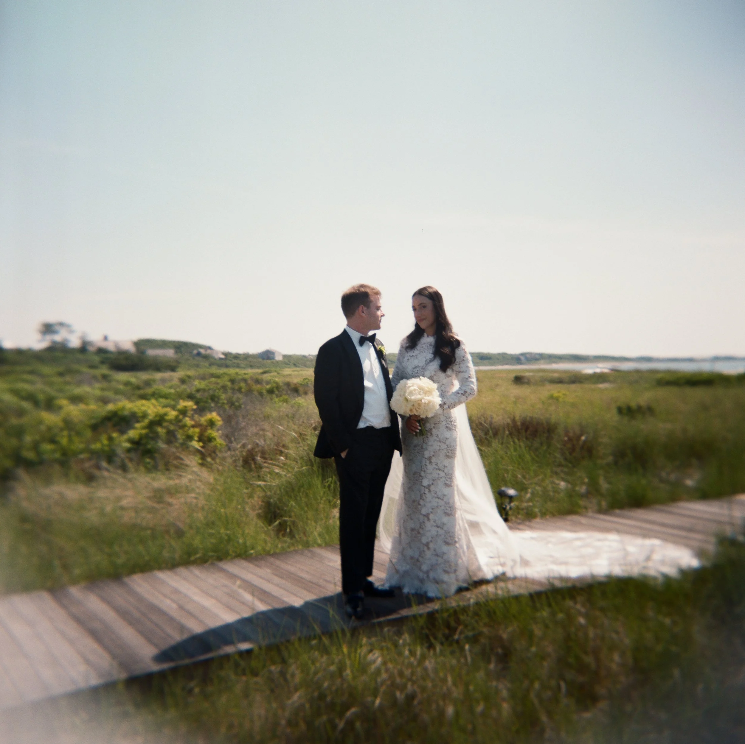 A bride and groom standing on a wooden walkway outdoors with green grass and bushes in the background, during a wedding photoshoot, under a clear sky.