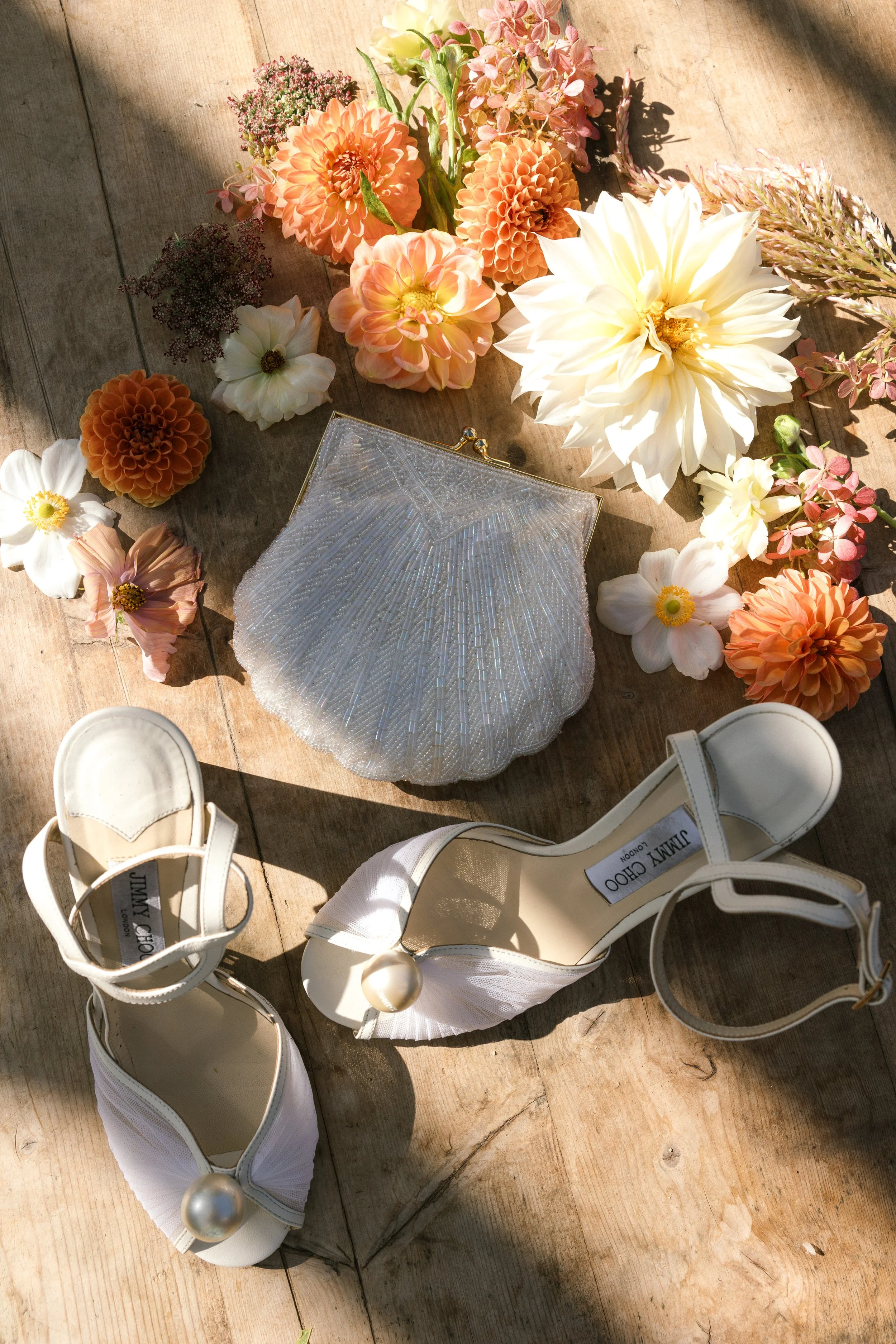 White high-heeled shoes with large pearl embellishments on a wooden surface surrounded by pink, white, and orange flowers, and a beaded clutch purse.
