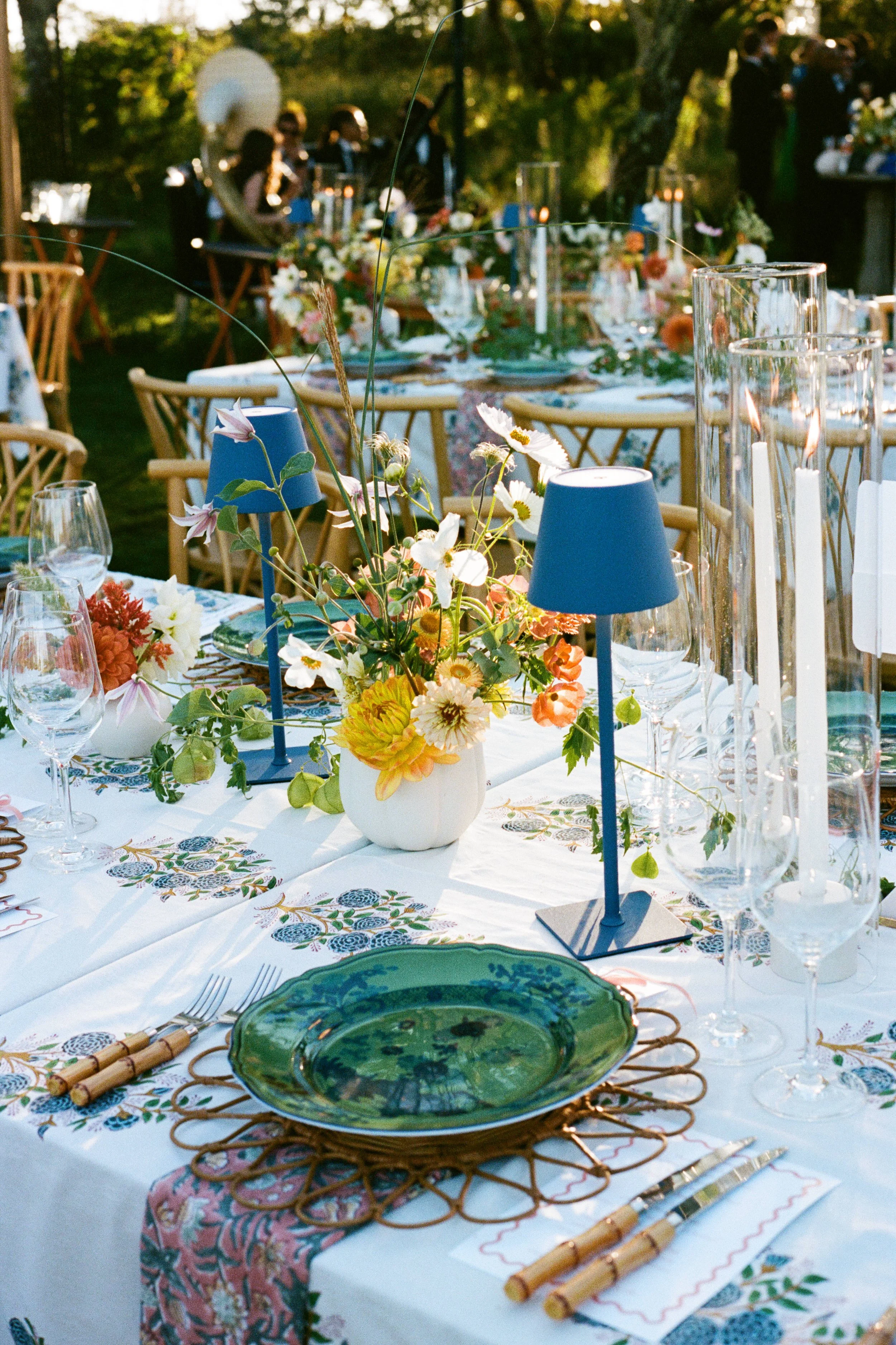 Elegantly set outdoor dining table with floral centerpiece, blue candle holders, glassware, and green plates, with a wooded background and guests in the distance.