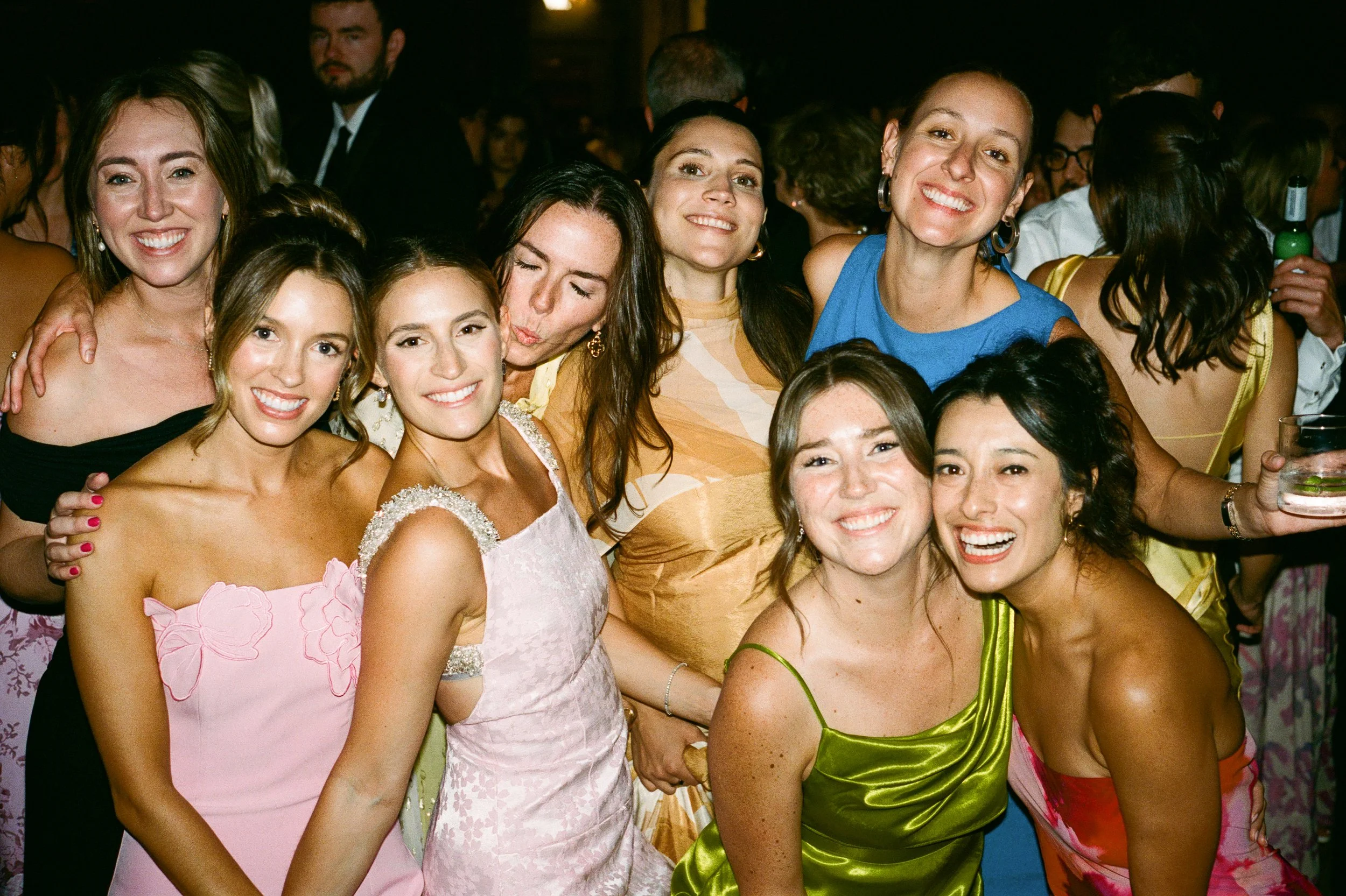 A group of women smiling and posing together at a celebration or party, dressed in colorful formal dresses.