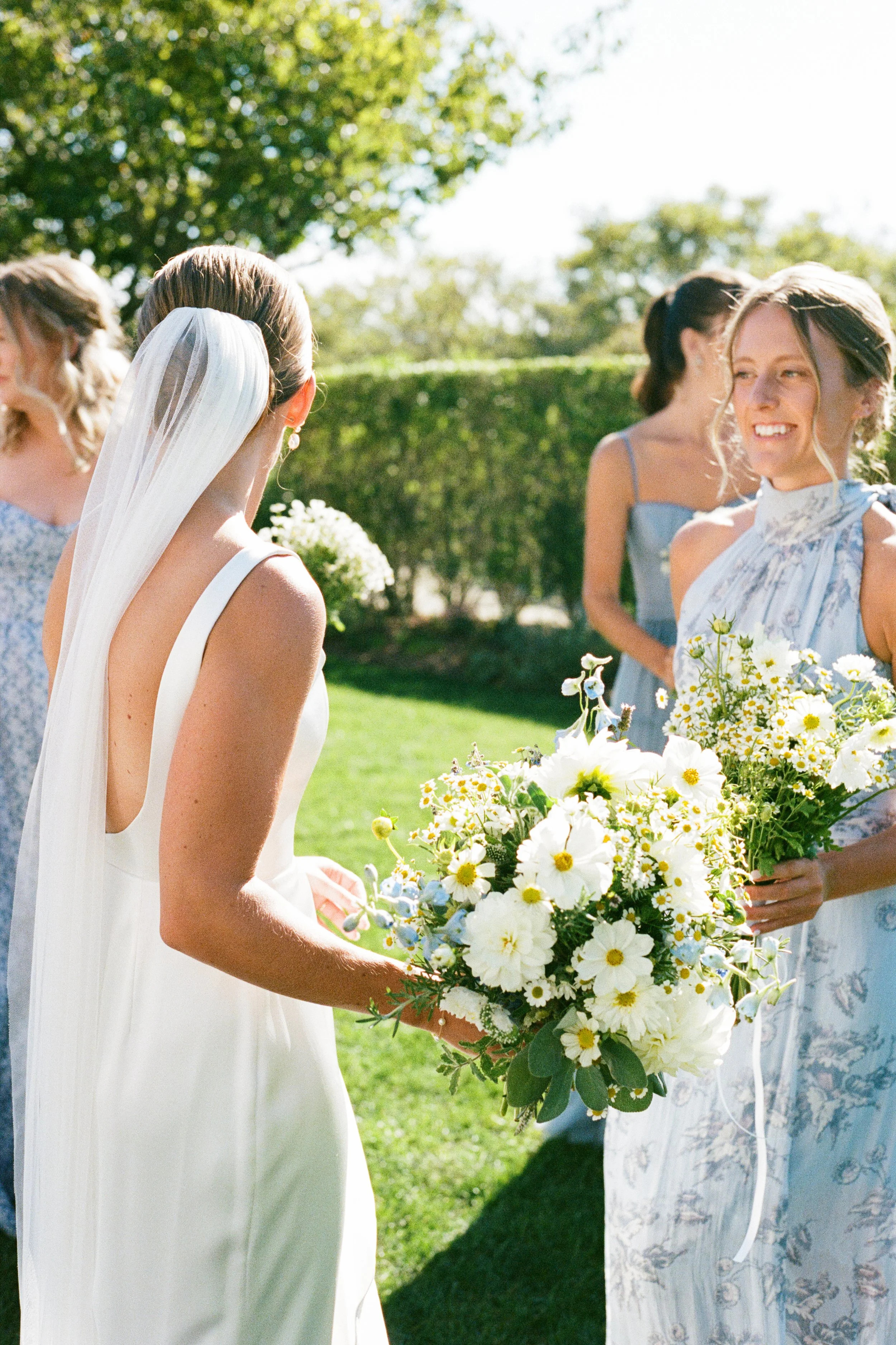 Bride in white wedding dress and veil holding a bouquet of white and yellow flowers talking to a woman in a light blue floral dress and holding a similar bouquet outdoors on a sunny day.