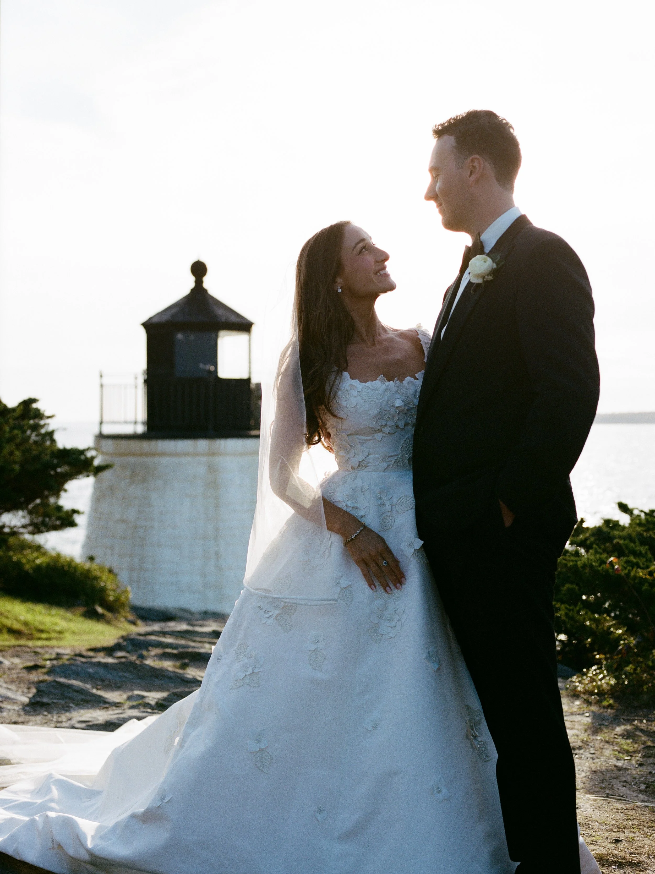 A bride and groom looking at each other, standing outdoors near a lighthouse during sunset.