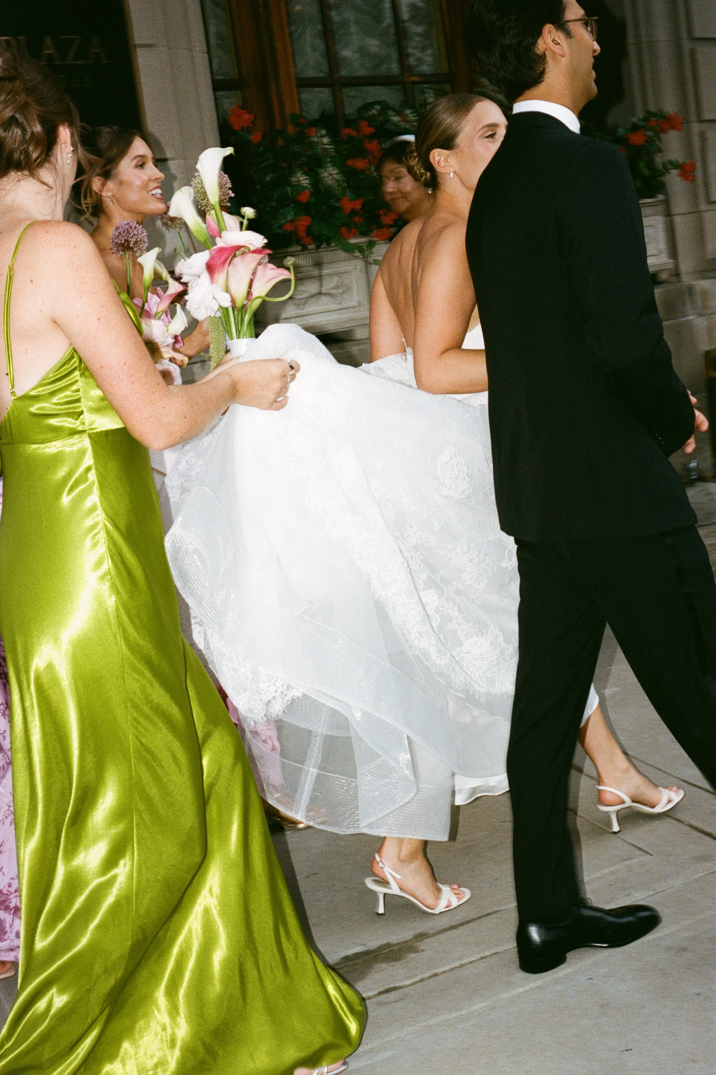 A bride in a white wedding dress, a man in a black tuxedo, and bridesmaids in colorful dresses, walking together during a wedding celebration.