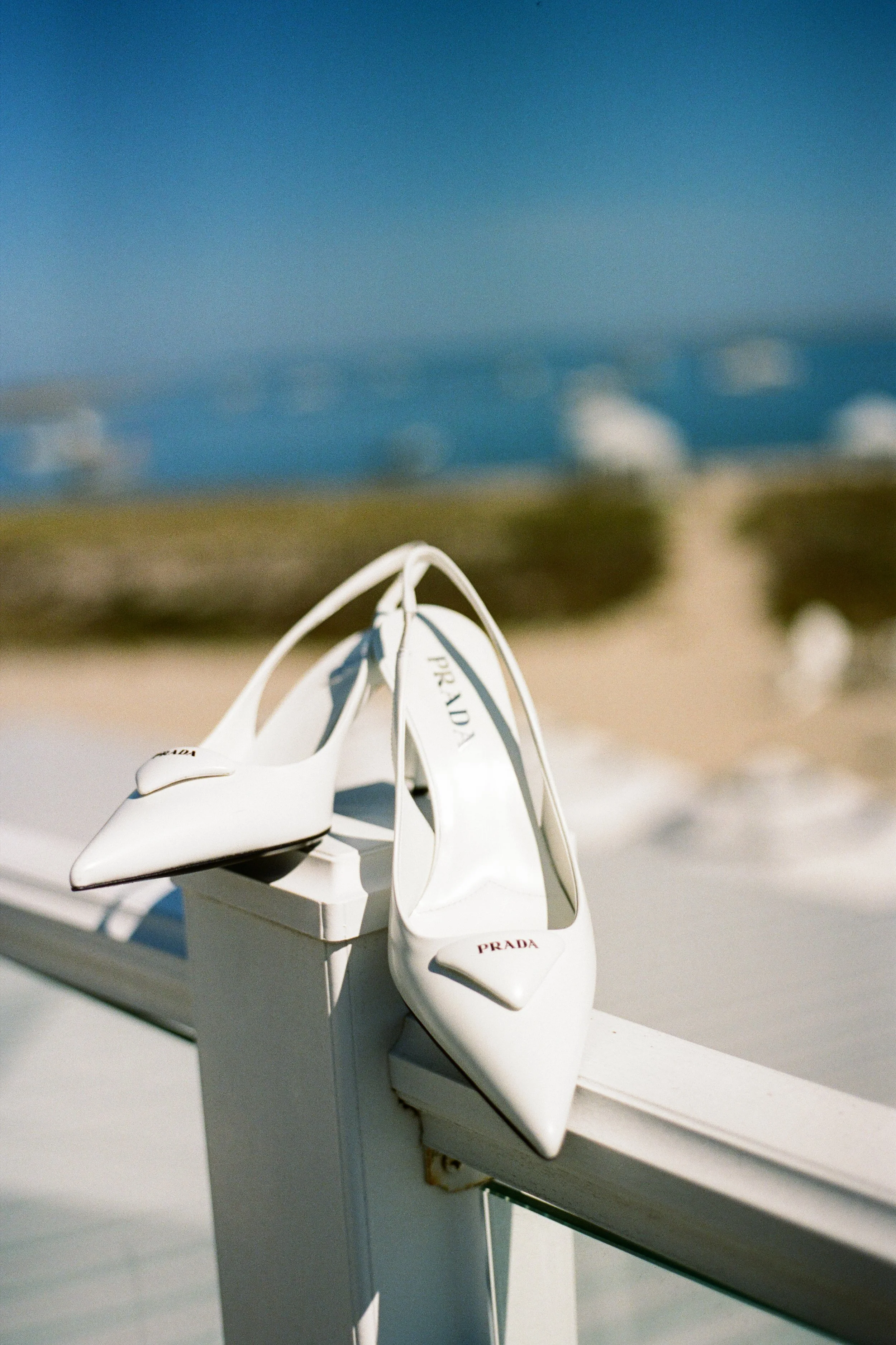 A pair of white Prada high-heeled shoes resting on a white railing with a blurred beach and ocean in the background.