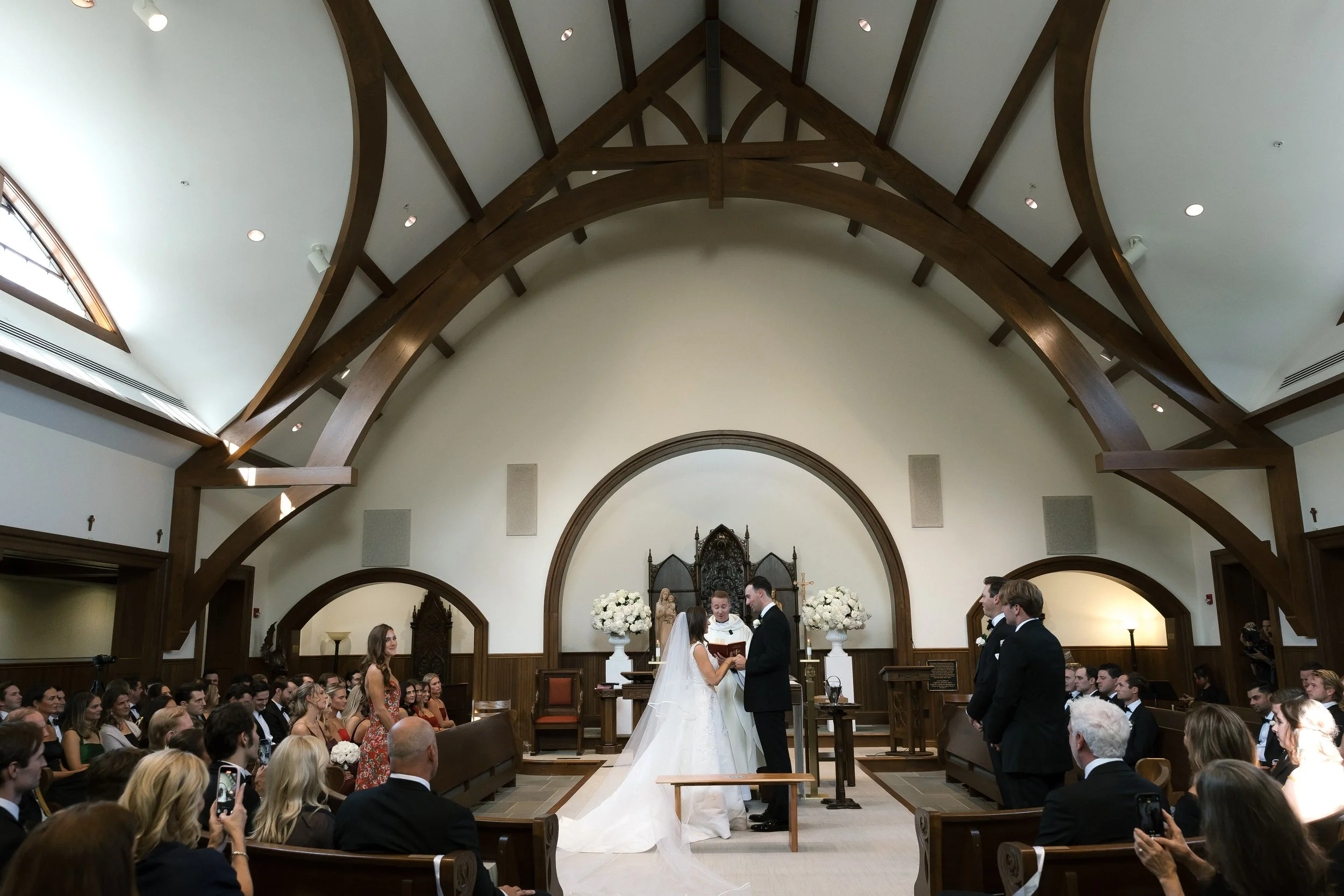 A wedding ceremony taking place inside a church with a bride and groom standing before an officiant. The church ceiling has large wooden beams and an altar decorated with white flowers. Guests are seated and watching the couple.