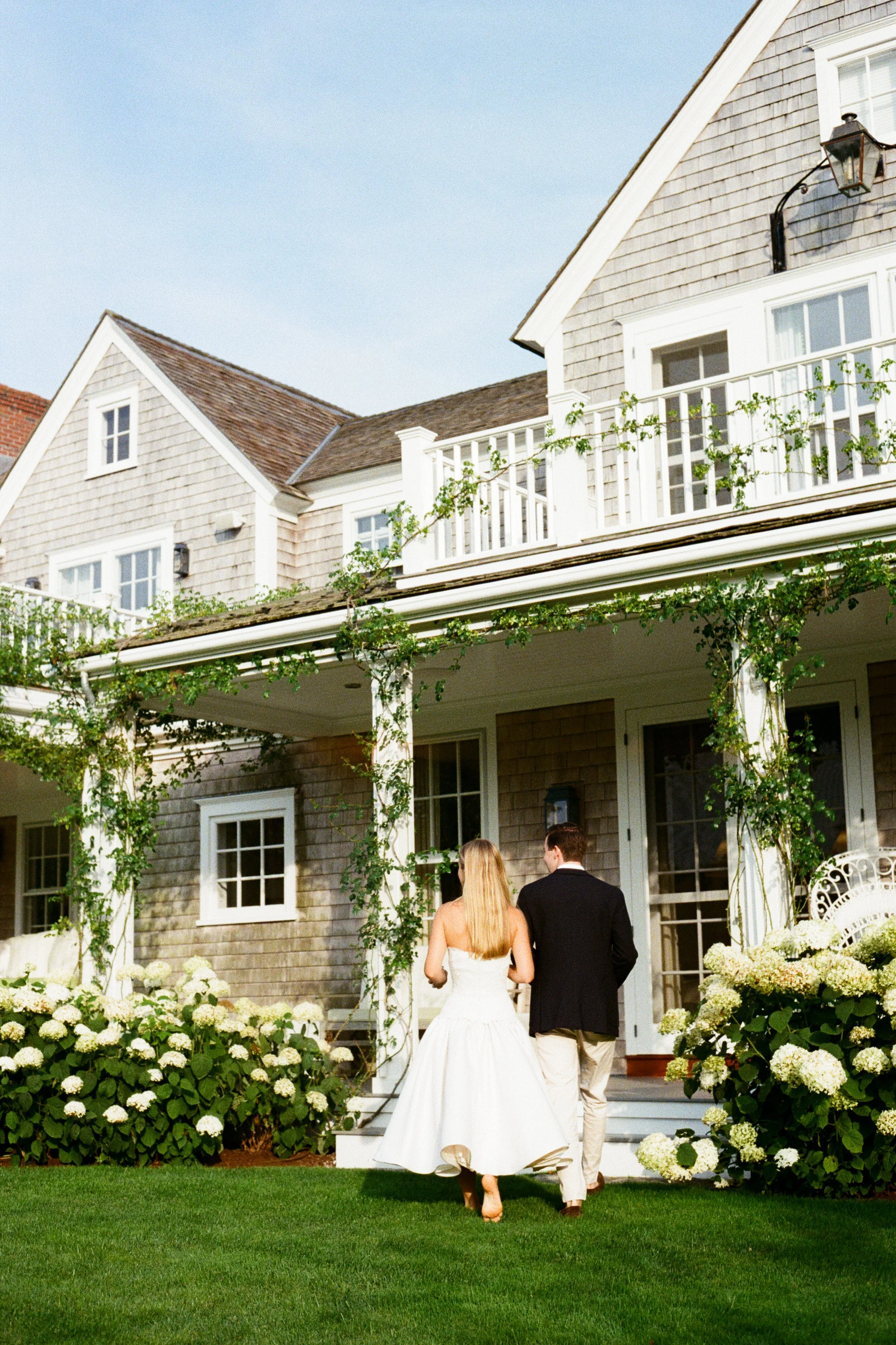 A couple dressed in wedding attire walking toward a house with a porch, surrounded by white hydrangeas outside.