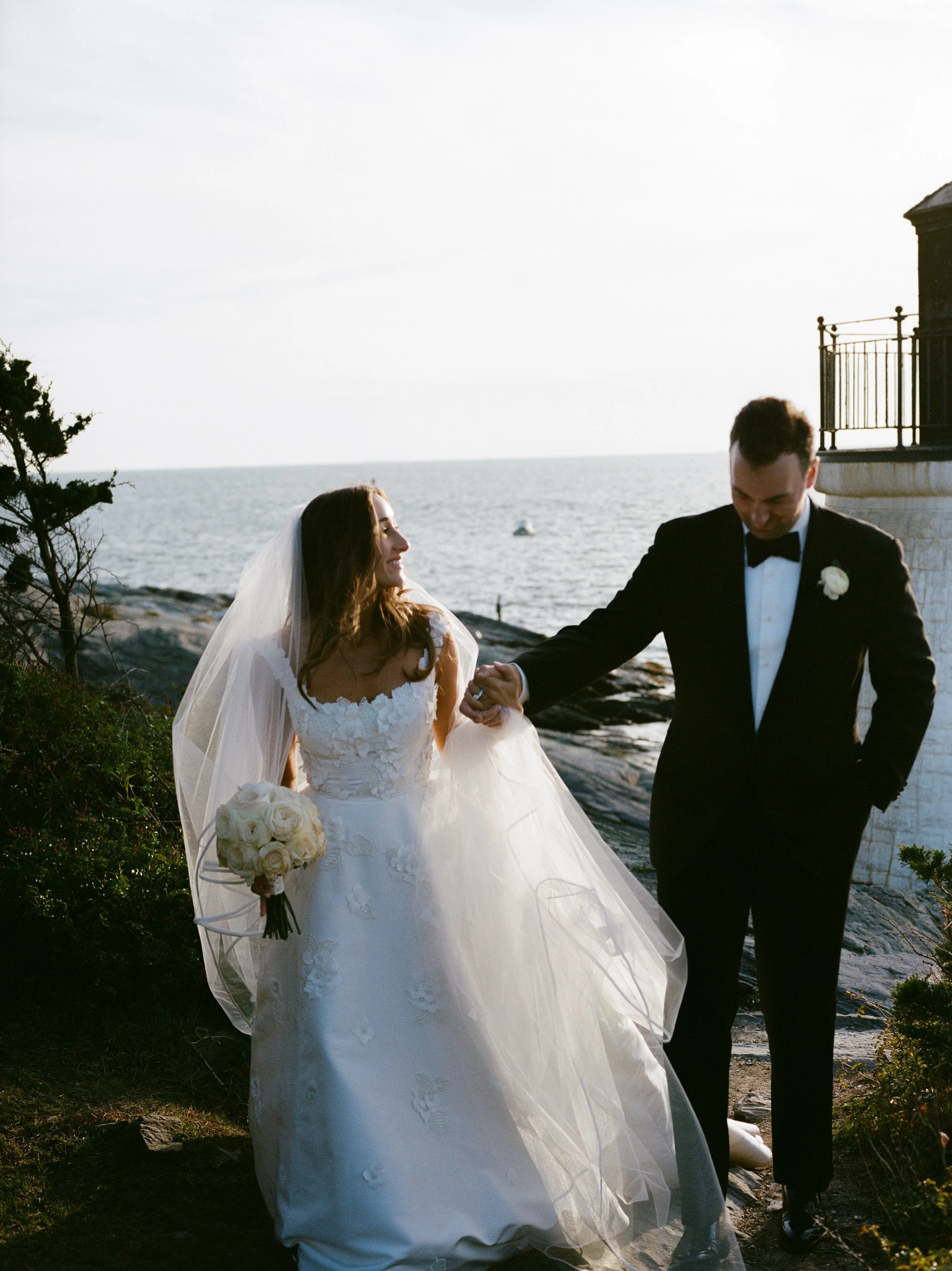 Bride and groom holding hands on a rocky shoreline during sunset, with the ocean and a small boat in the background.