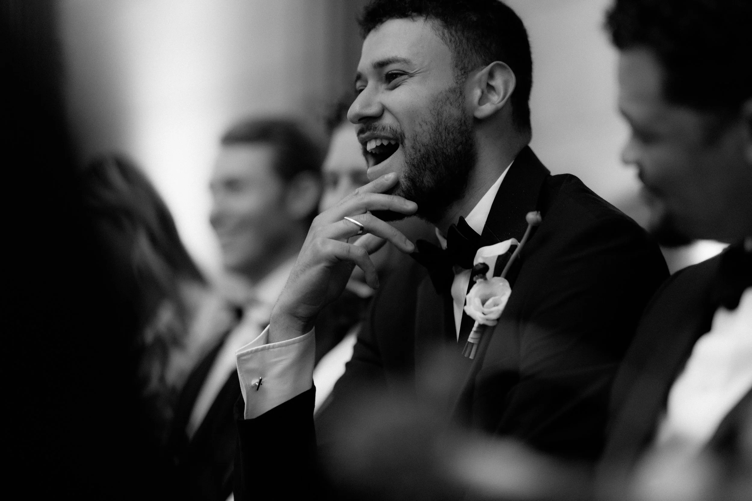 A group of men dressed in tuxedos, seated at a formal event, with one man in focus smiling and touching his chin.