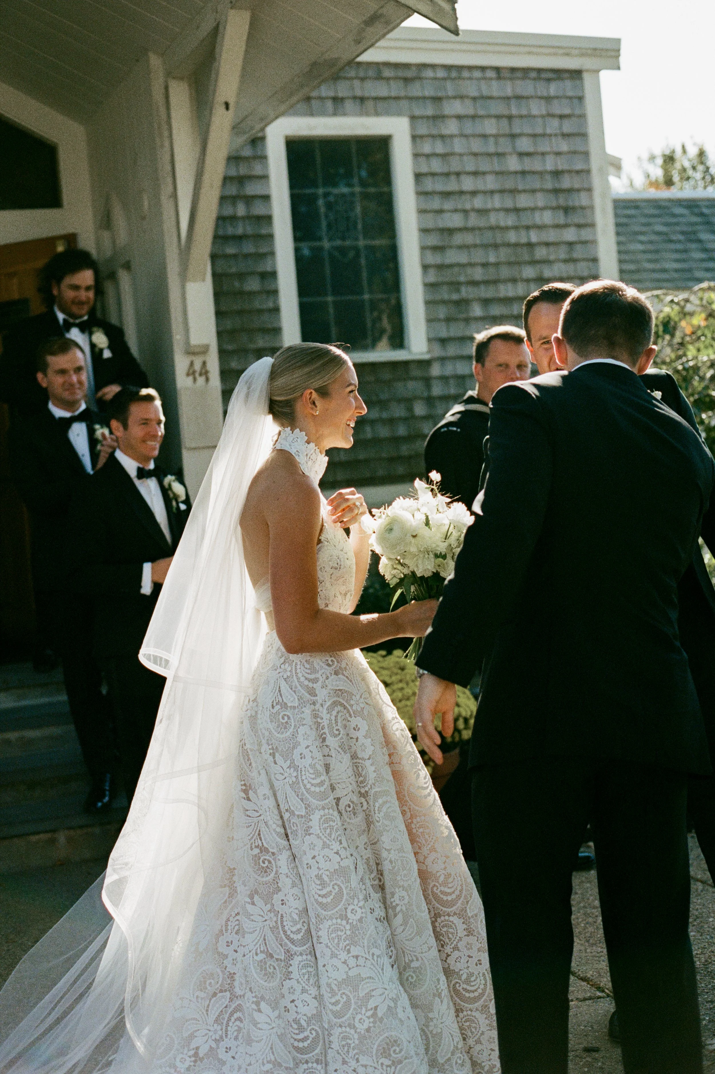 Bride in a lace wedding gown holding a bouquet, smiling, as she interacts with her groom outside a house. Wedding party members, dressed in black tuxedos, stand nearby.