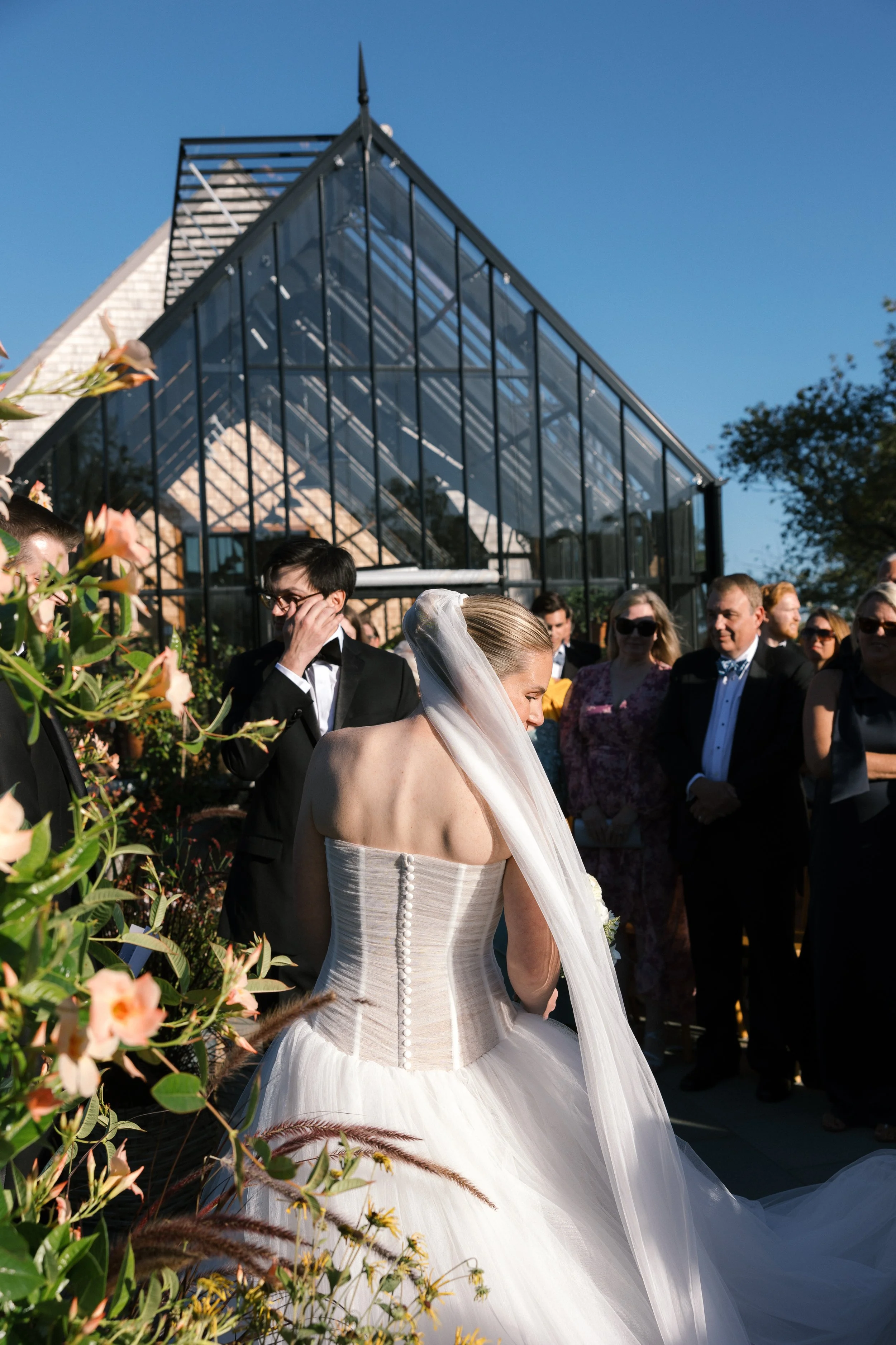 Bride in a white wedding dress and veil is surrounded by wedding guests outdoors near a glass greenhouse on a sunny day.