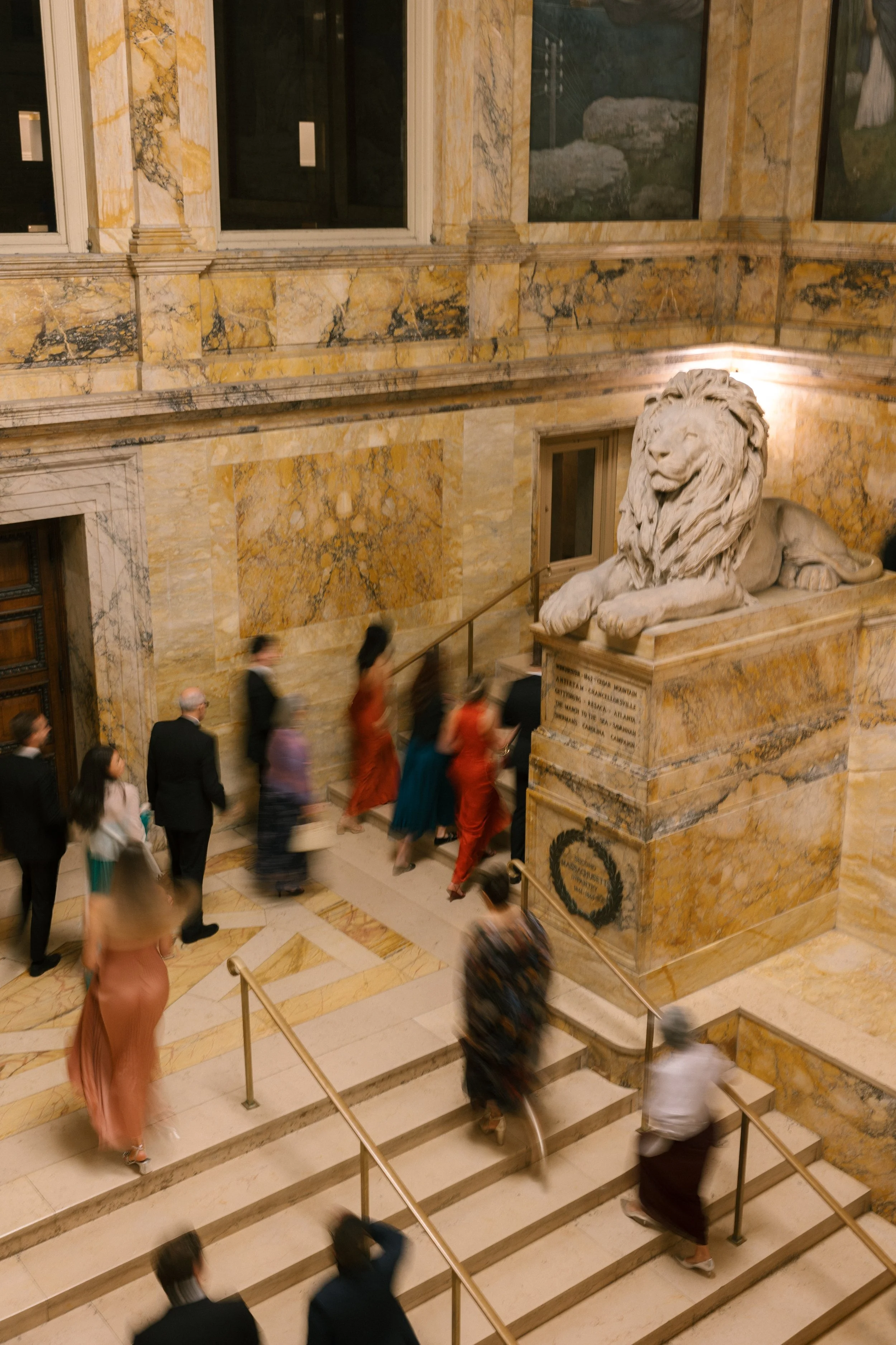 People ascending marble stairs in a grand, ornate building with marble walls and a large stone lion sculpture at the landing.