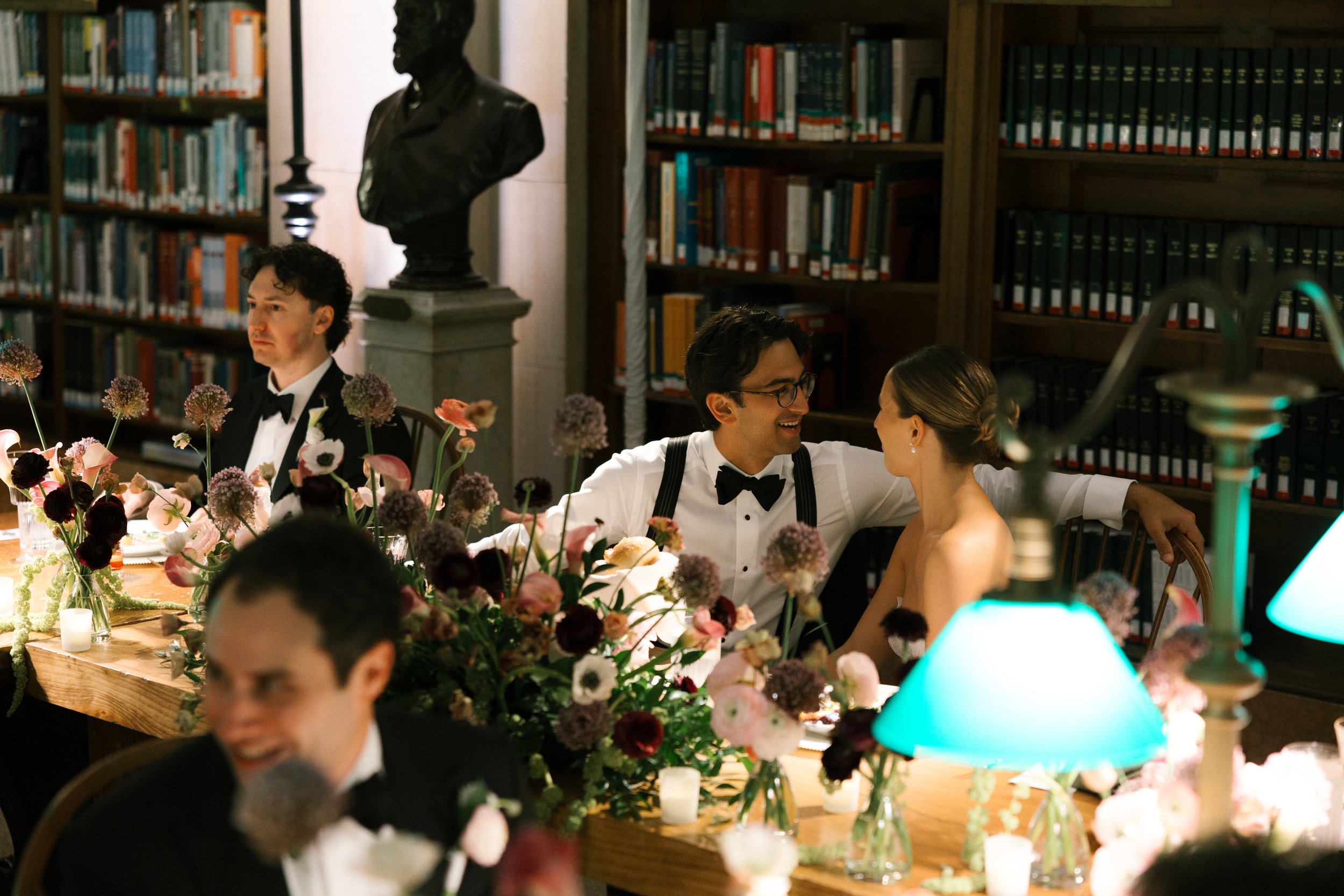 People in formal attire at a dinner party in a room with bookshelves and a bust sculpture, decorated with flowers and candles.