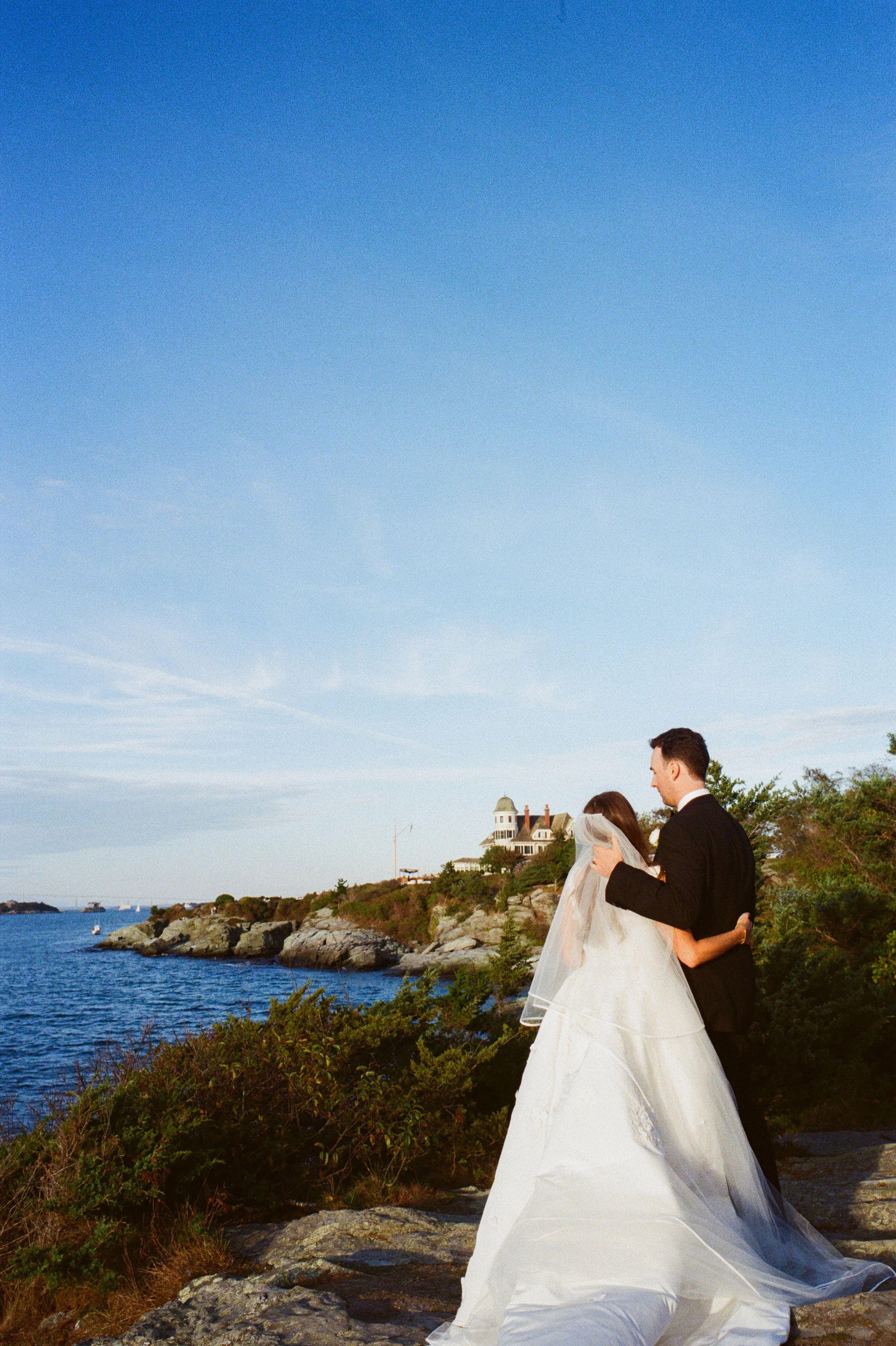 A bride and groom in wedding attire embrace on a rocky coastline with a house and trees in the background, under a clear blue sky.