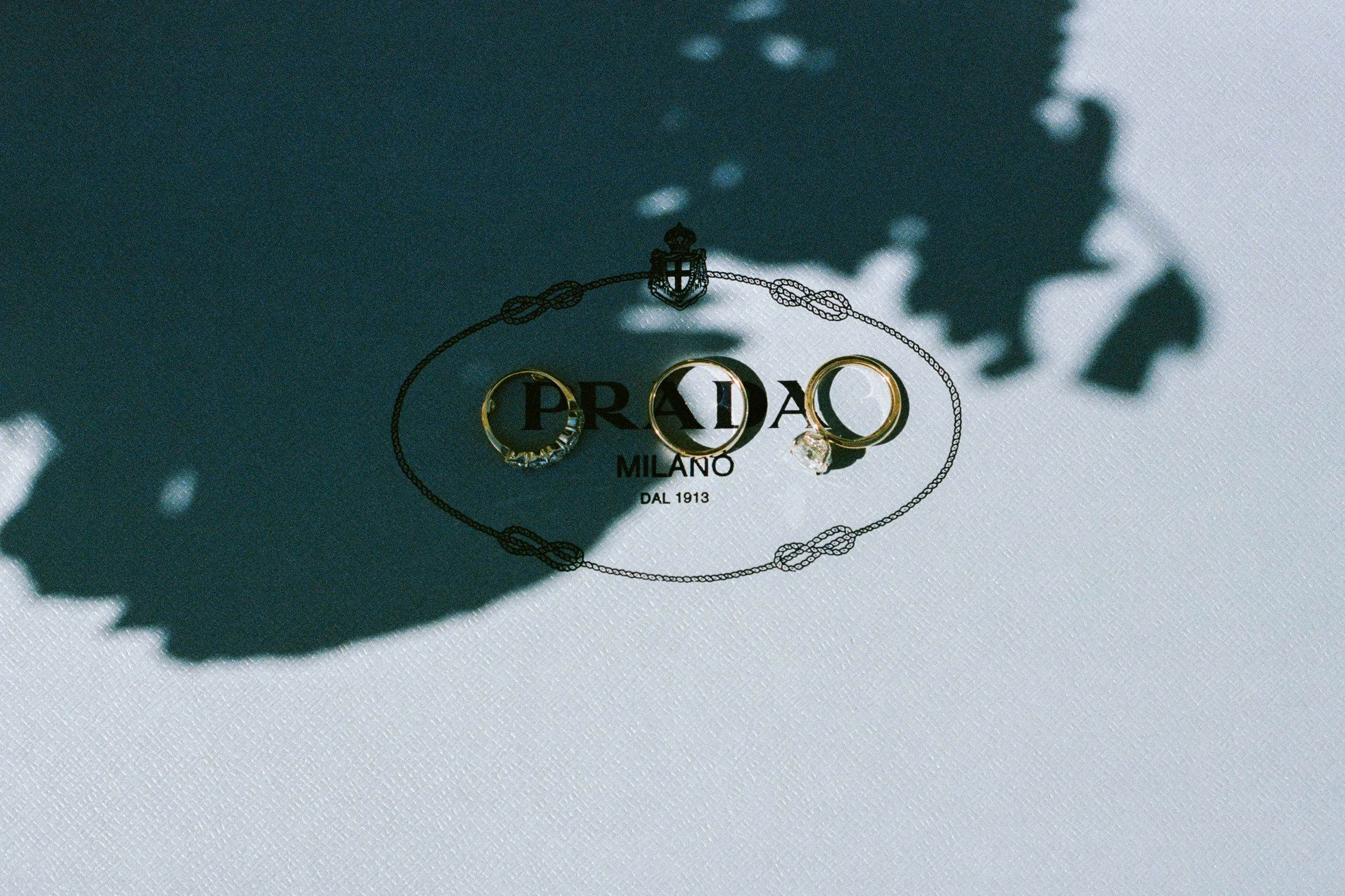 Prada jewelry box with rings and a chain, casting shadows on a white textured surface.