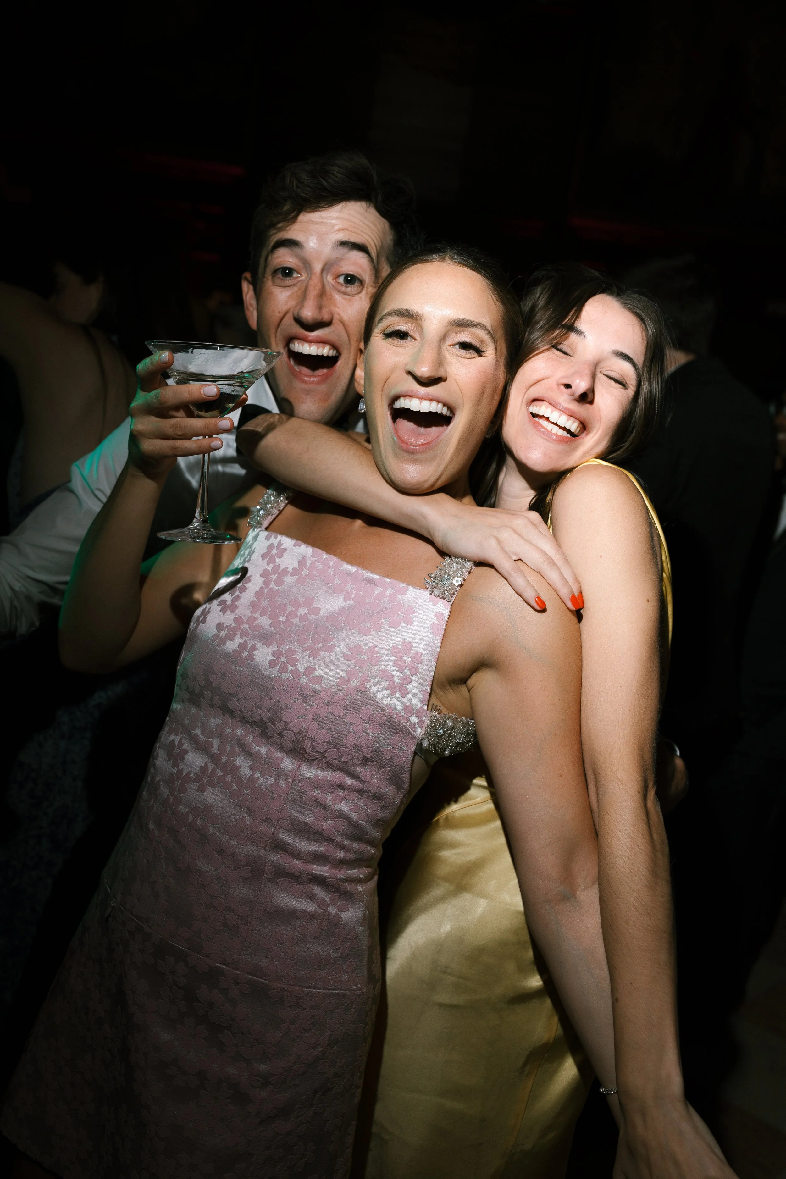Three friends hugging and smiling at a celebration or party, one holding a cocktail glass, wearing formal dresses.